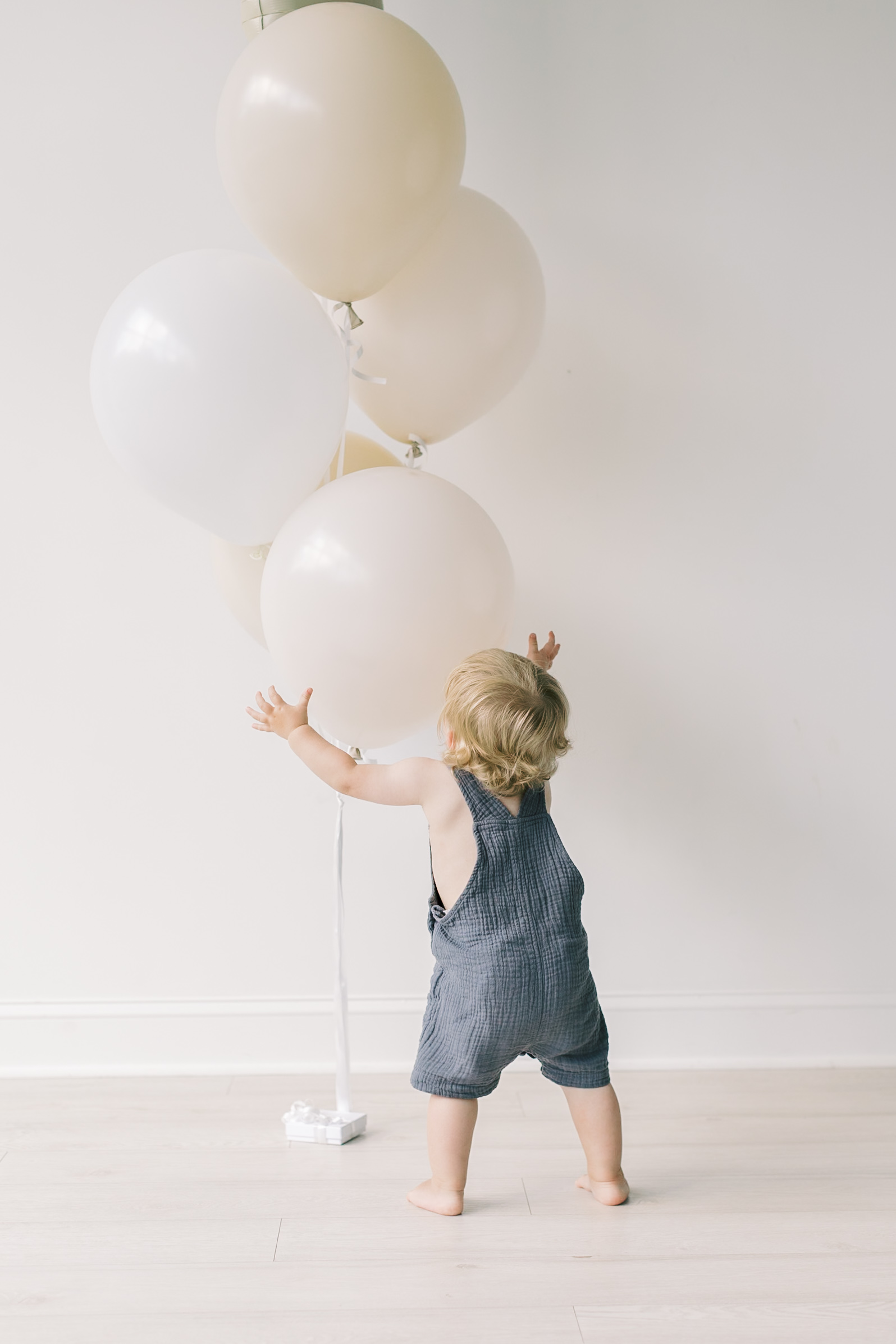 A blonde toddler in blue overalls chases some white balloons in a studio after exploring spots for a kid's birthday party in raleigh, NC