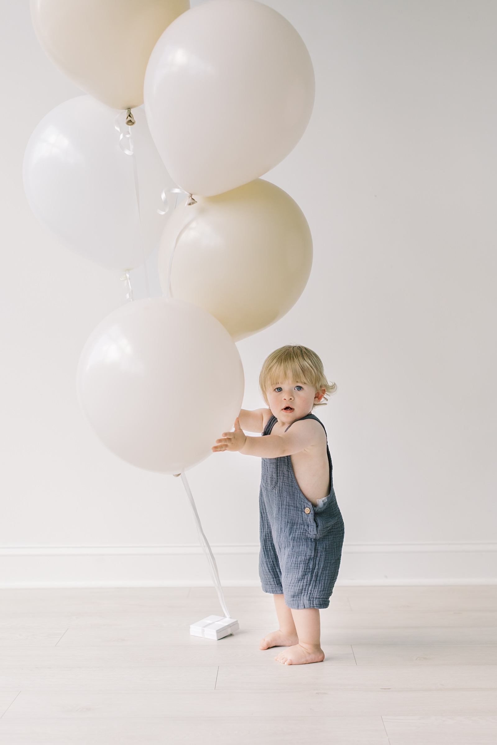 A toddler in blue overalls plays with big white balloons in a studio before his kid's birthday party in raleigh, NC
