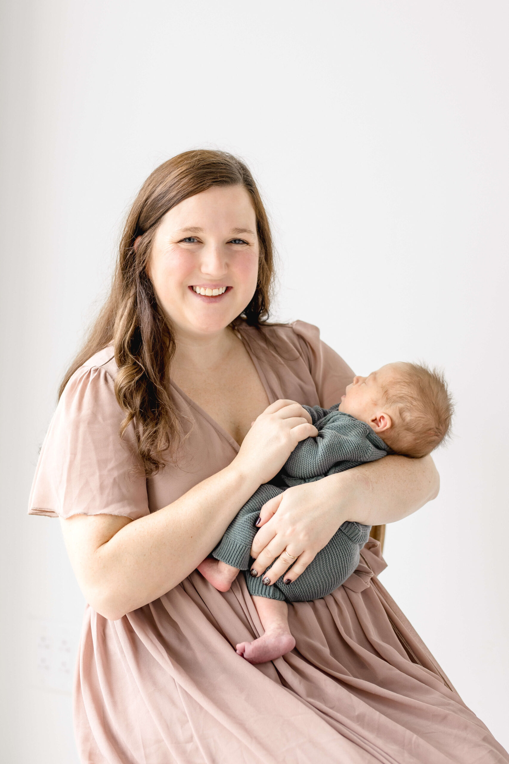 A happy new mom in a pink dress holds her sleeping newborn baby in her arms while sitting on a stool in a studio