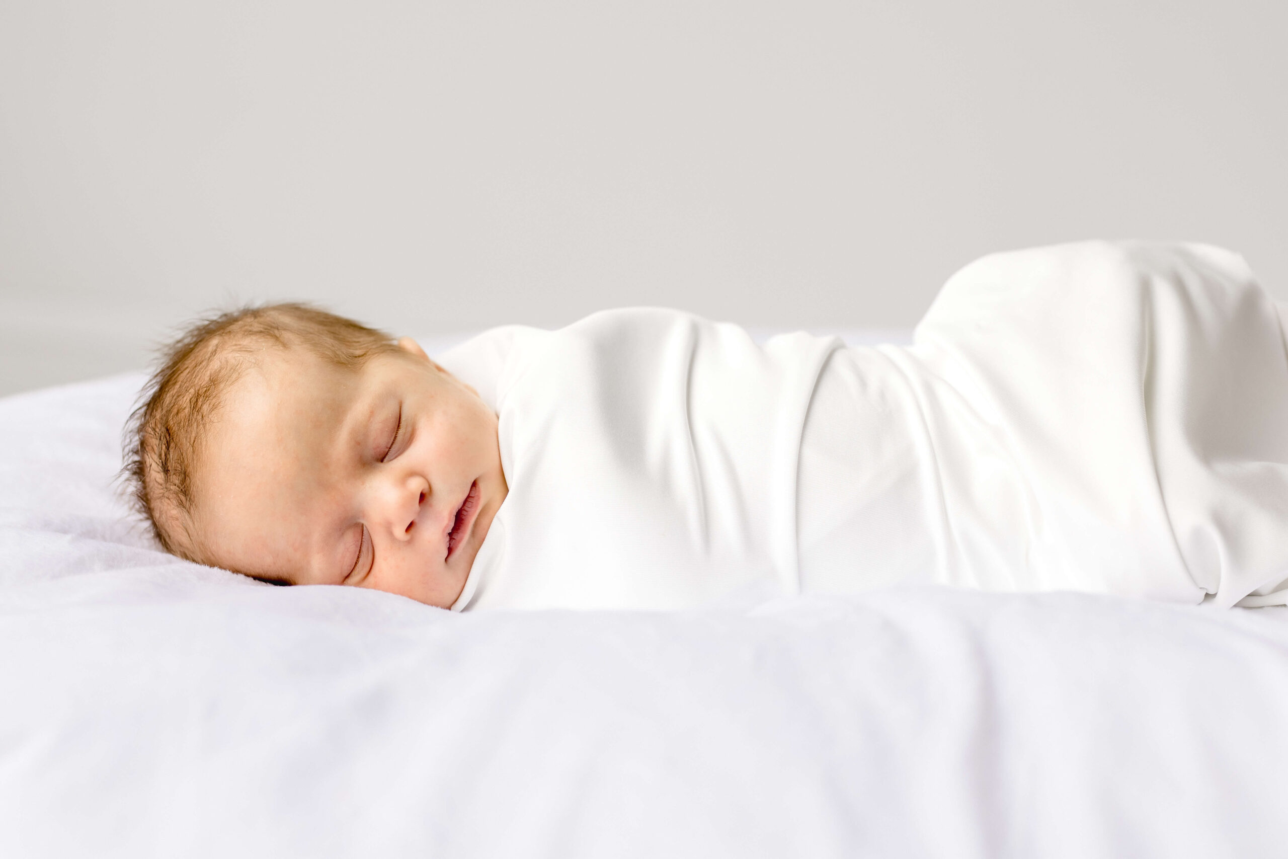 A newborn baby with red hair sleeps in a white swaddle on its side