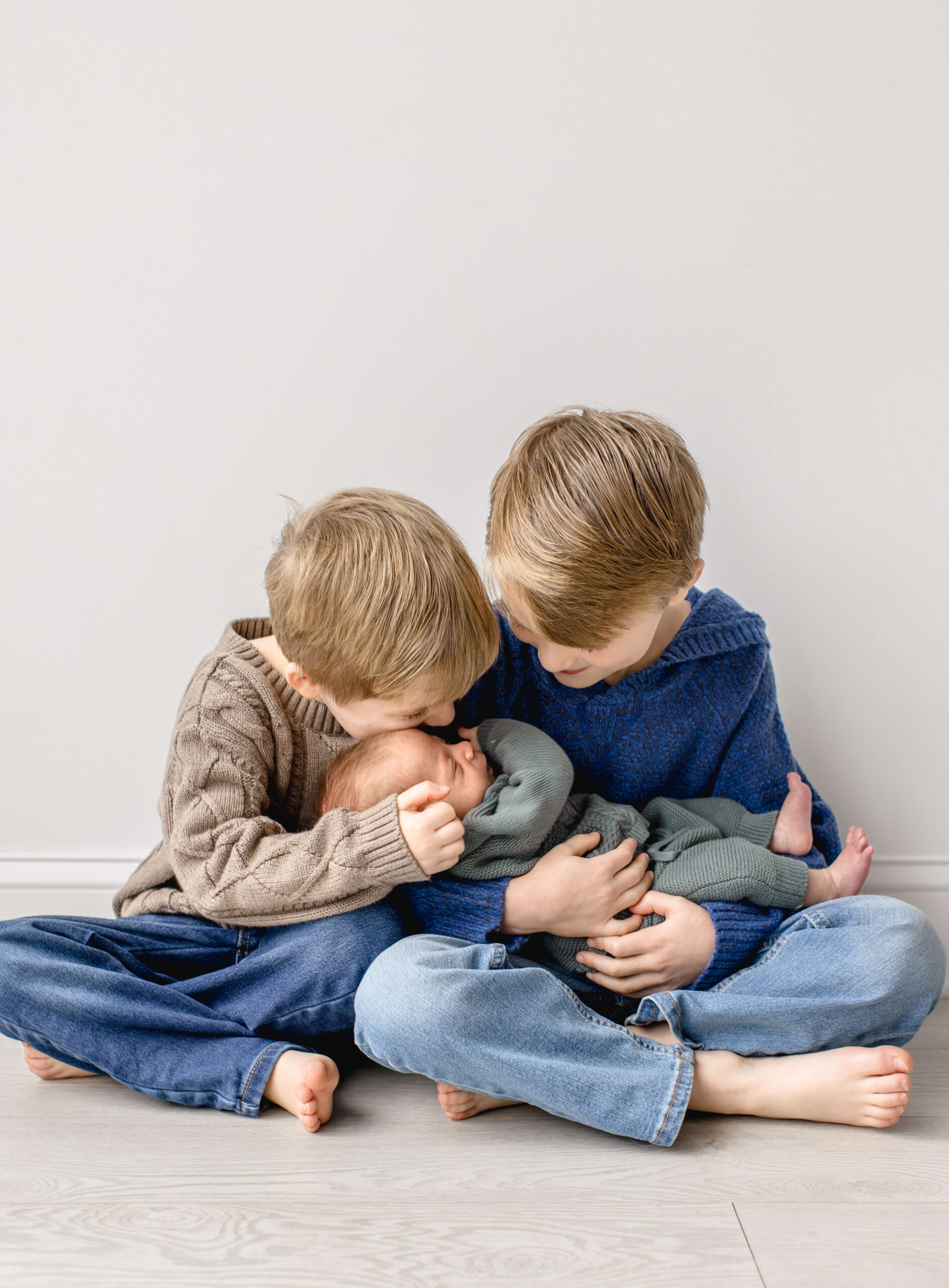 A newborn baby in a green knit onesie laying big brothers' laps as one leans down to kiss him in a studio after visiting a pediatric chiropractors in raleigh