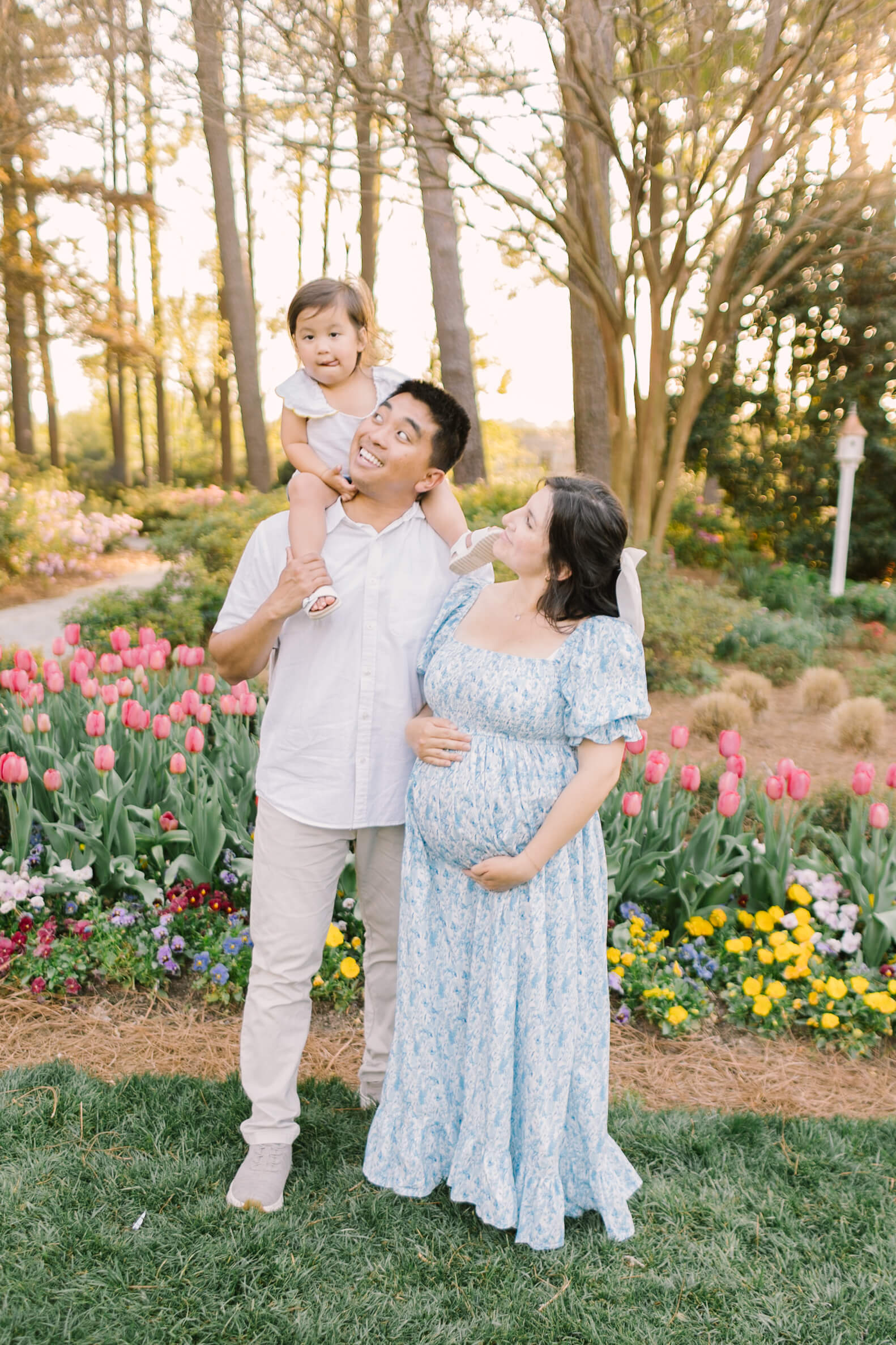 A toddler girl sits on dad's shoulders while mom and dad smile up to her in a flower garden