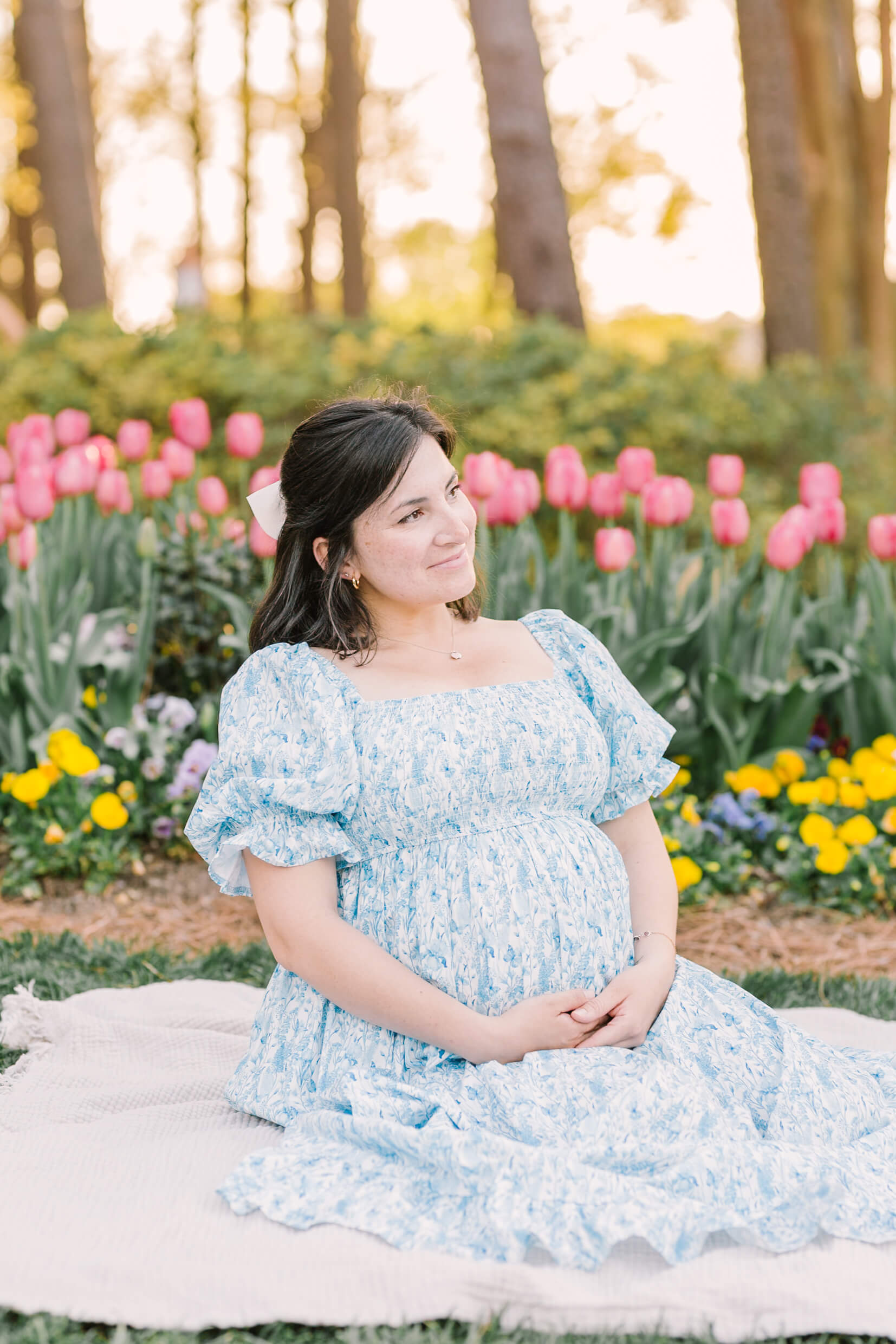 A happy mother to be sits on a picnic blanket in a pink tulip garden at sunset in a blue maternity dress