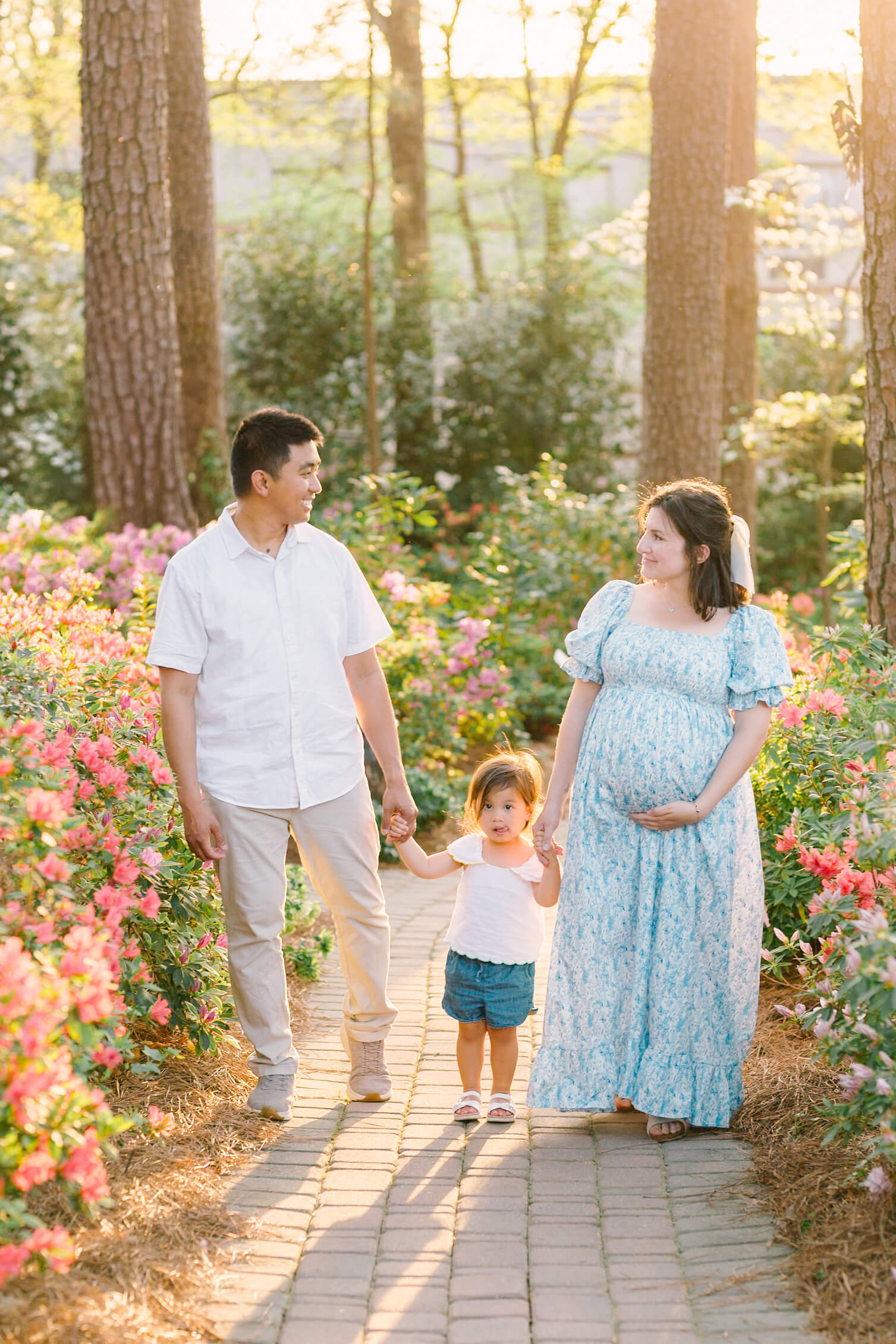 Expecting parents walk in a vibrant garden path at sunset smiling at each other and holding hands with their toddler daughter after a relaxing prenatal massage in raleigh