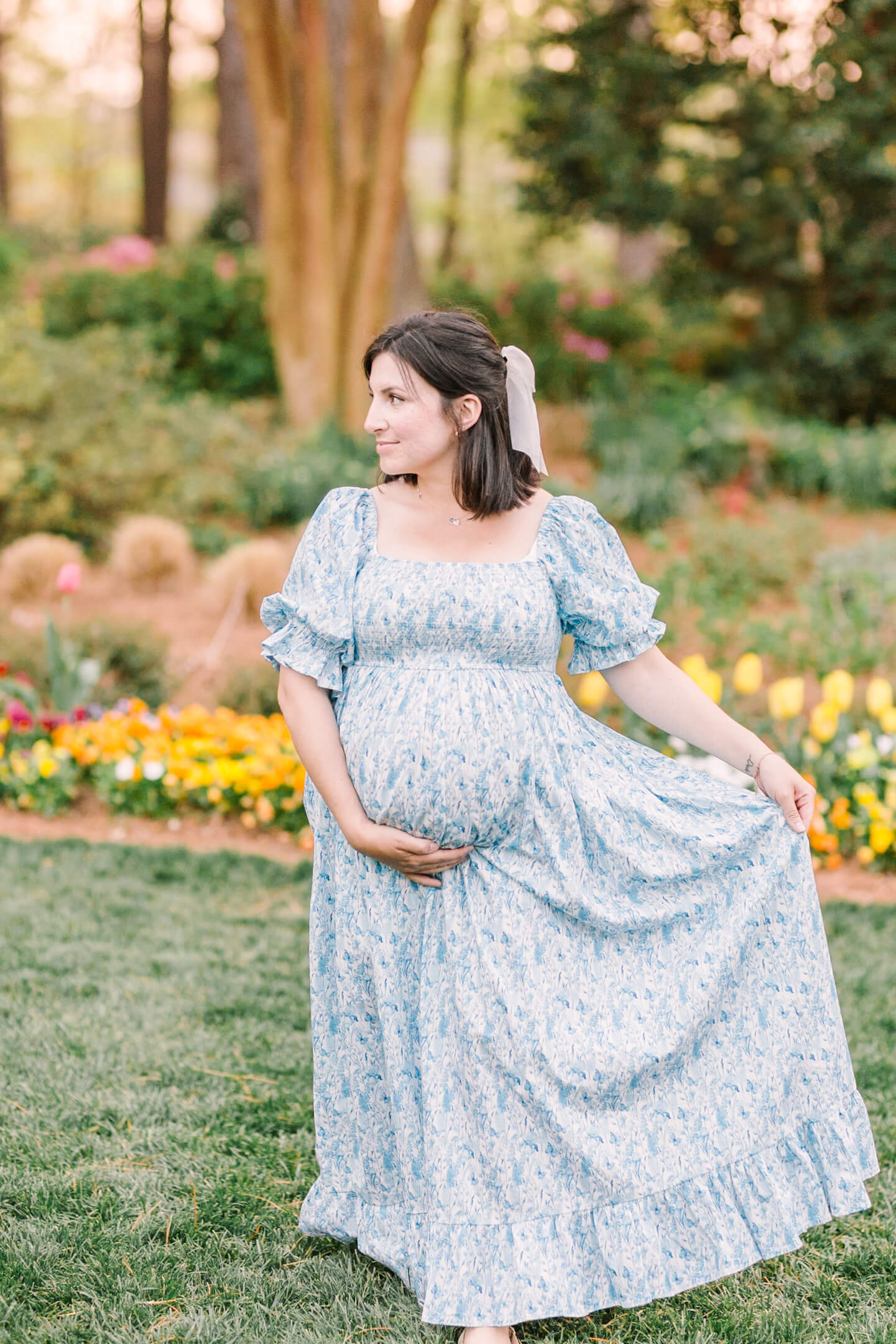 A smiling pregnant woman walks in a flower garden lawn playing with her blue floral print maternity dress after a prenatal massage in raleigh