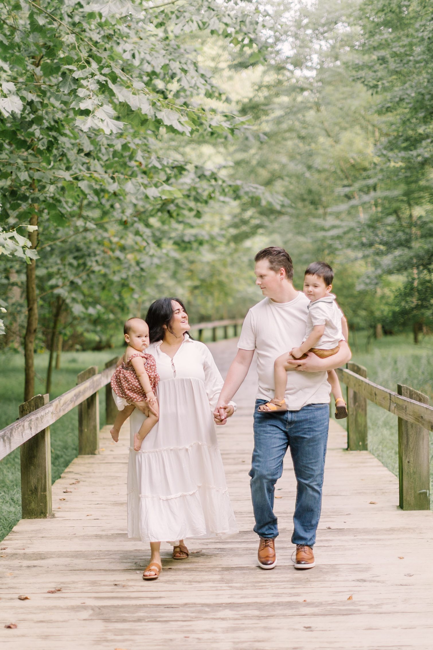 Happy mom and dad in white walk on a boardwalk holding hands and carrying their two toddlers after Easter brunch in raleigh