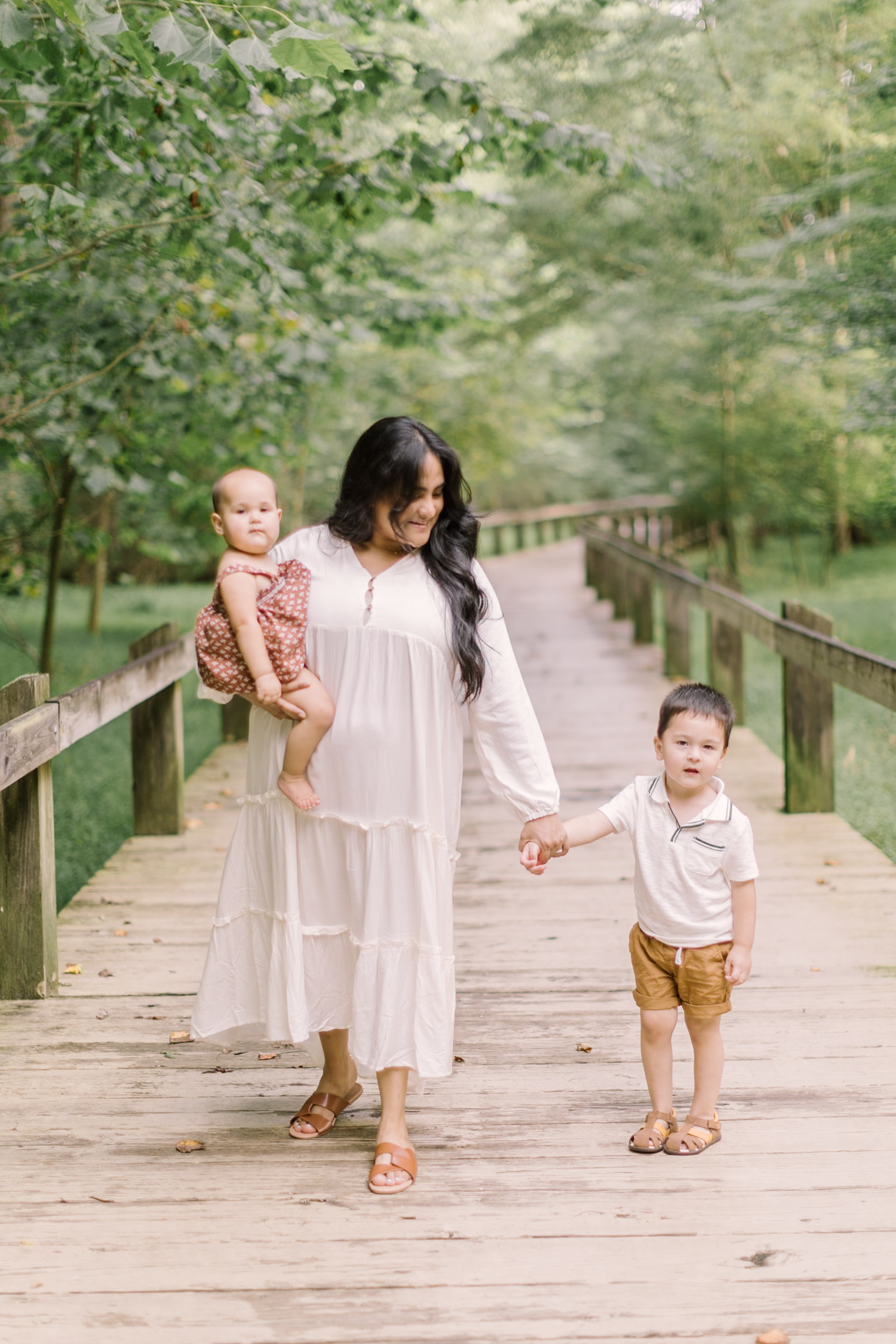 A happy mom in a white dress walks with her toddler son and baby daughter on her hip on a boardwalk