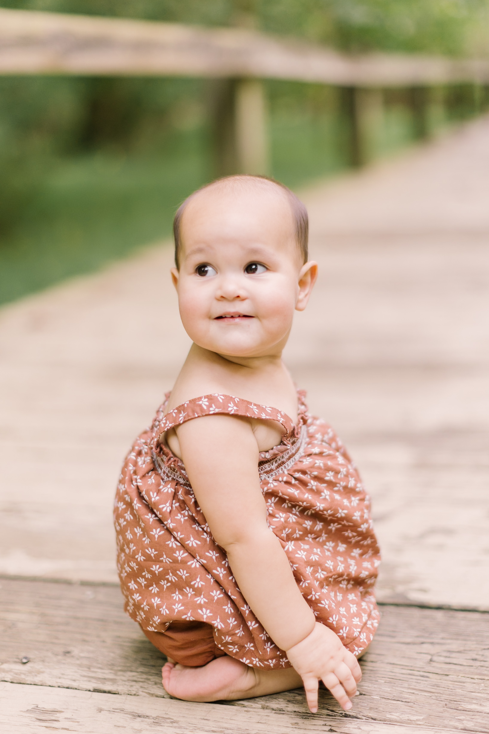 A baby girl smiles over her shoulder while playing on a boardwalk in a pink dress before Easter brunch in raleigh