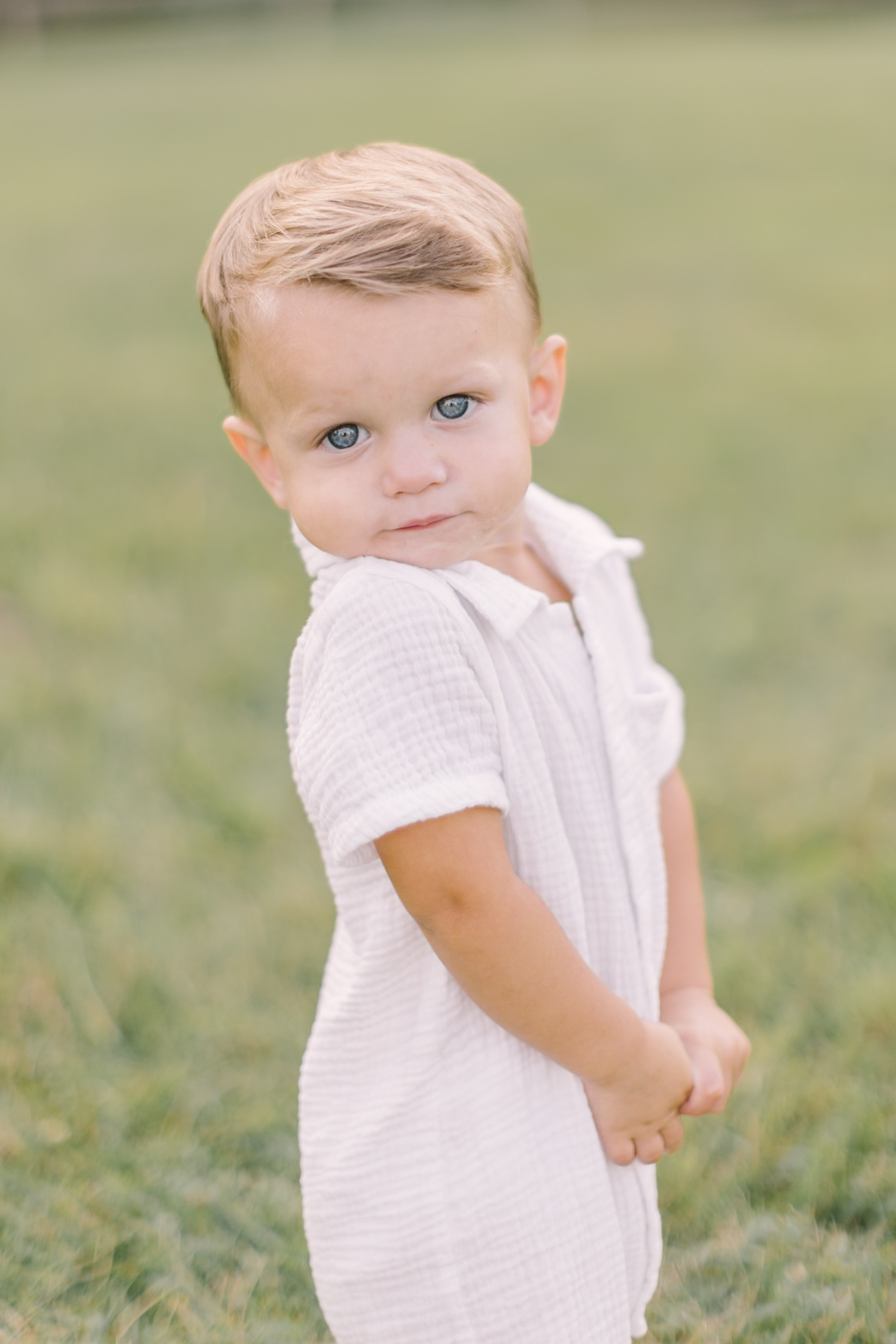 A toddler boy in a white romper smiles over his shoulder while walking in a lawn