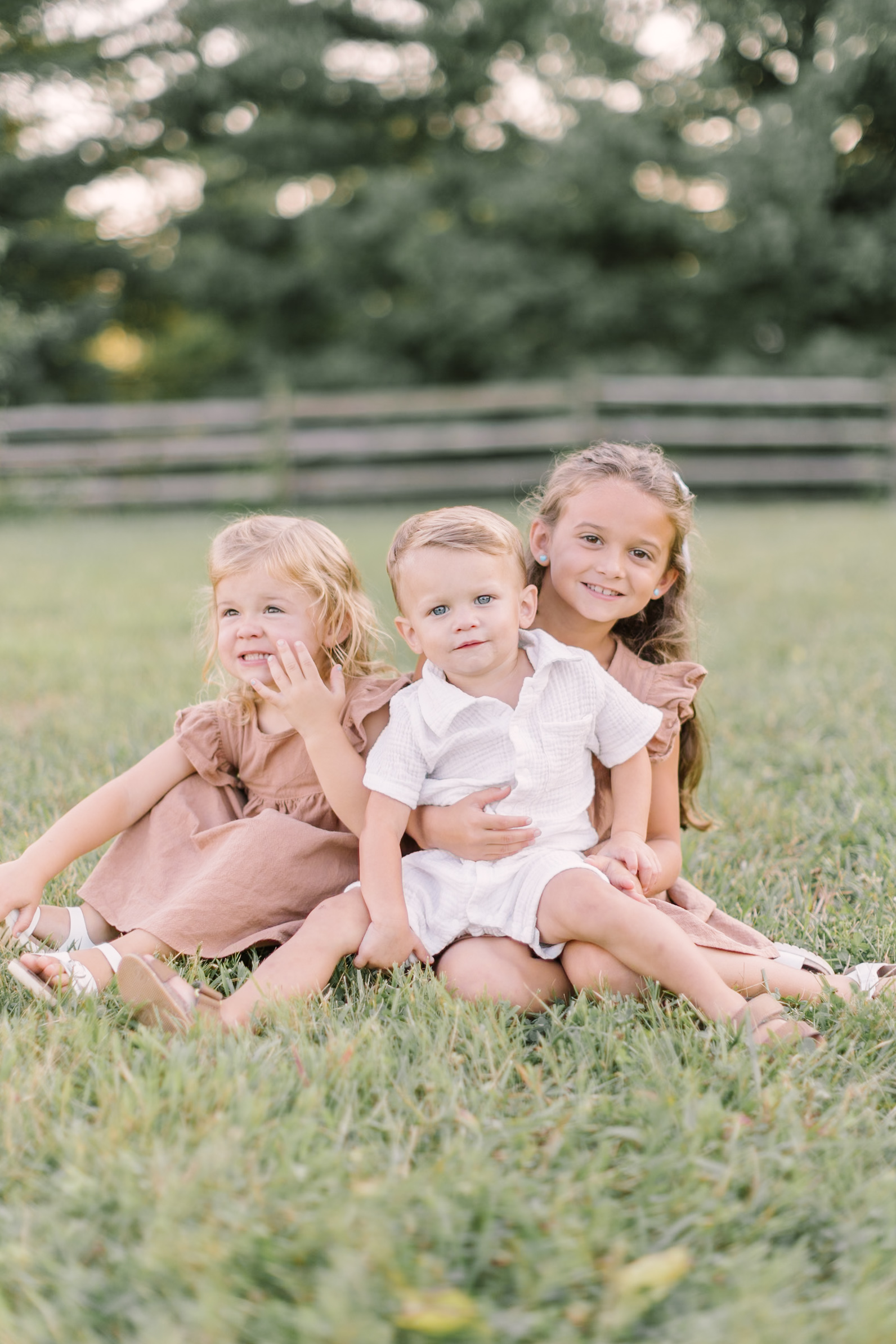 Happy toddler sisters in matching pink dresses sit in a pasture at sunset with their baby brother in white in their laps after visiting easter egg hunts in Raleigh, NC