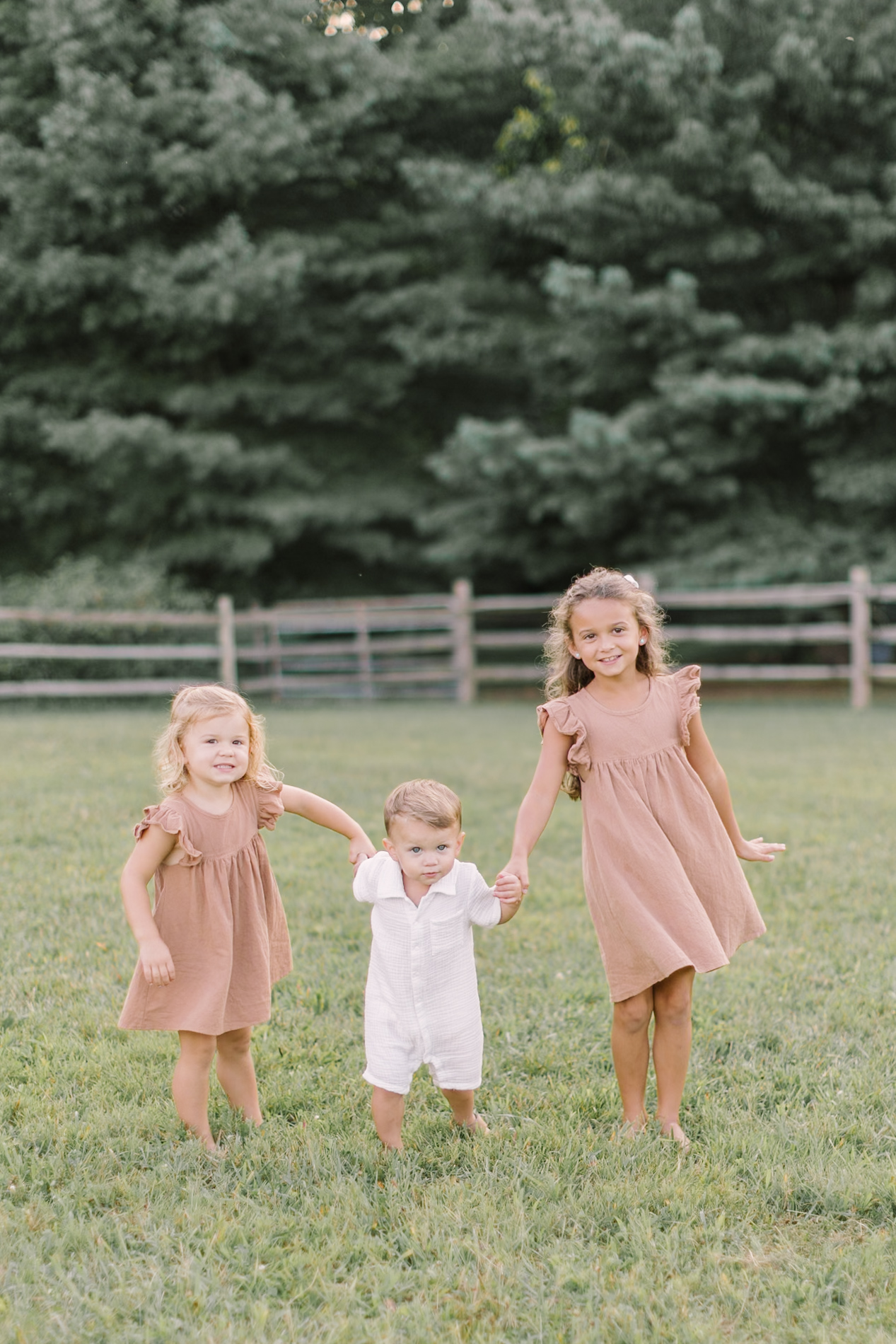 Toddler sisters in matching pink dresses walk their baby brother through a pasture in a white romper before heading to easter egg hunts in Raleigh, NC