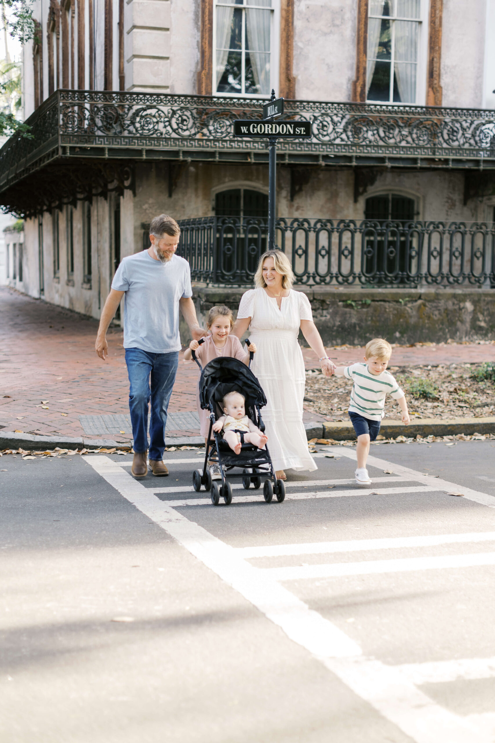 A mom and dad walk on a crosswalk with their three toddlers holding their hands and pushing baby carriage