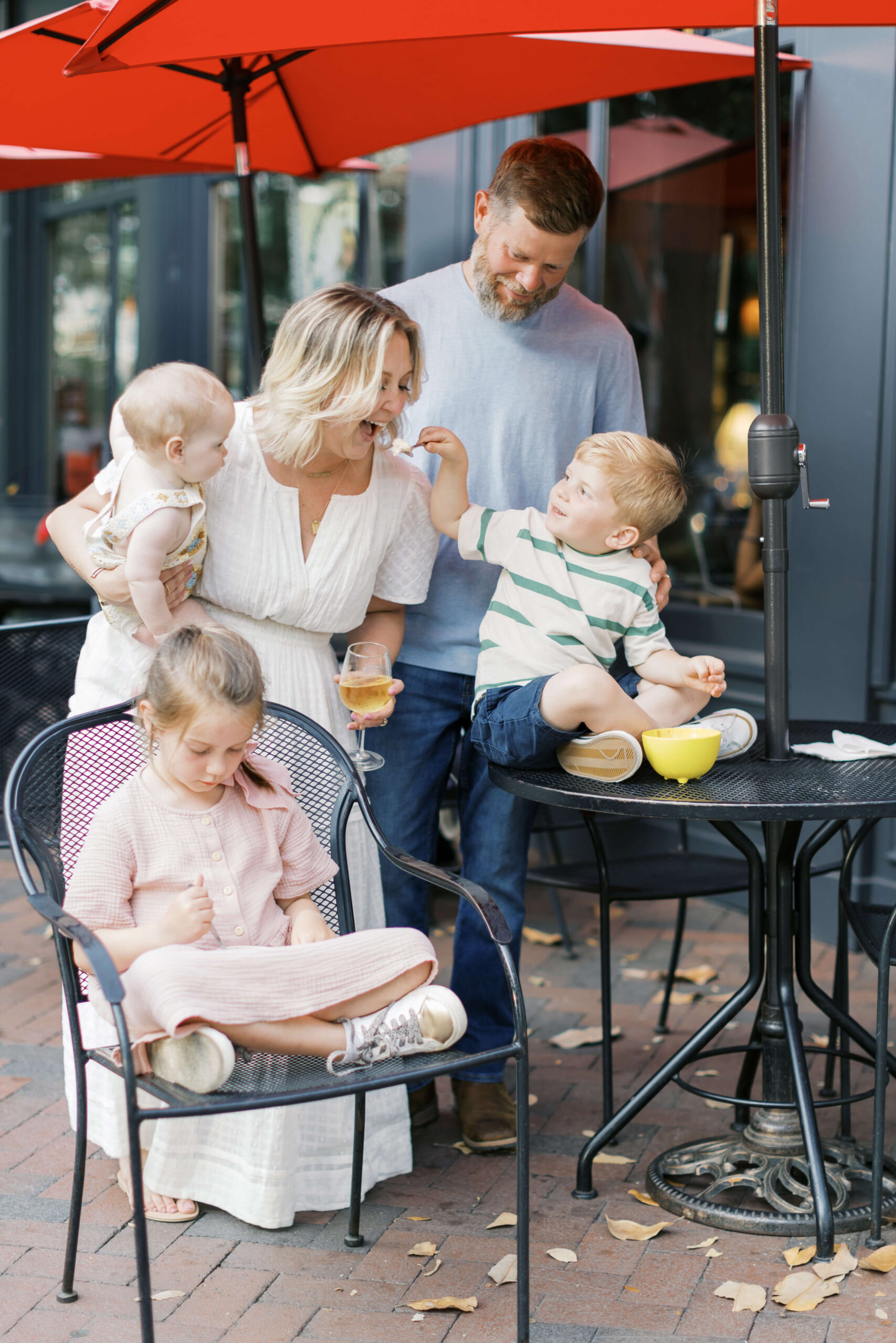 A toddler boy sit on a patio table feeding his mom from a bowl while at one of the family-friendly restaurants in Raleigh, NC with dad and two sisters