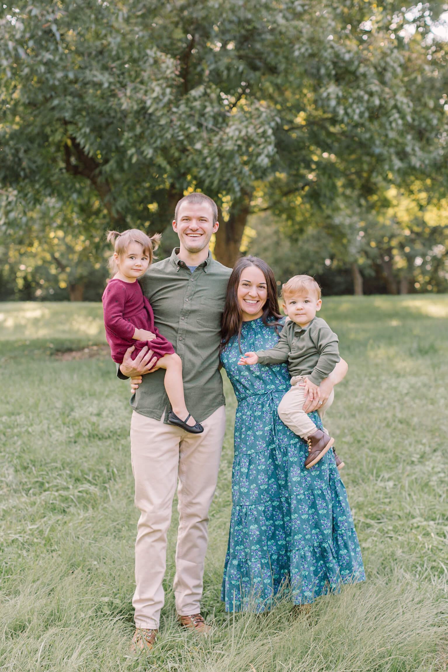 Smiling mom and dad stand in a park lawn holding their toddler son and daughter on their hips