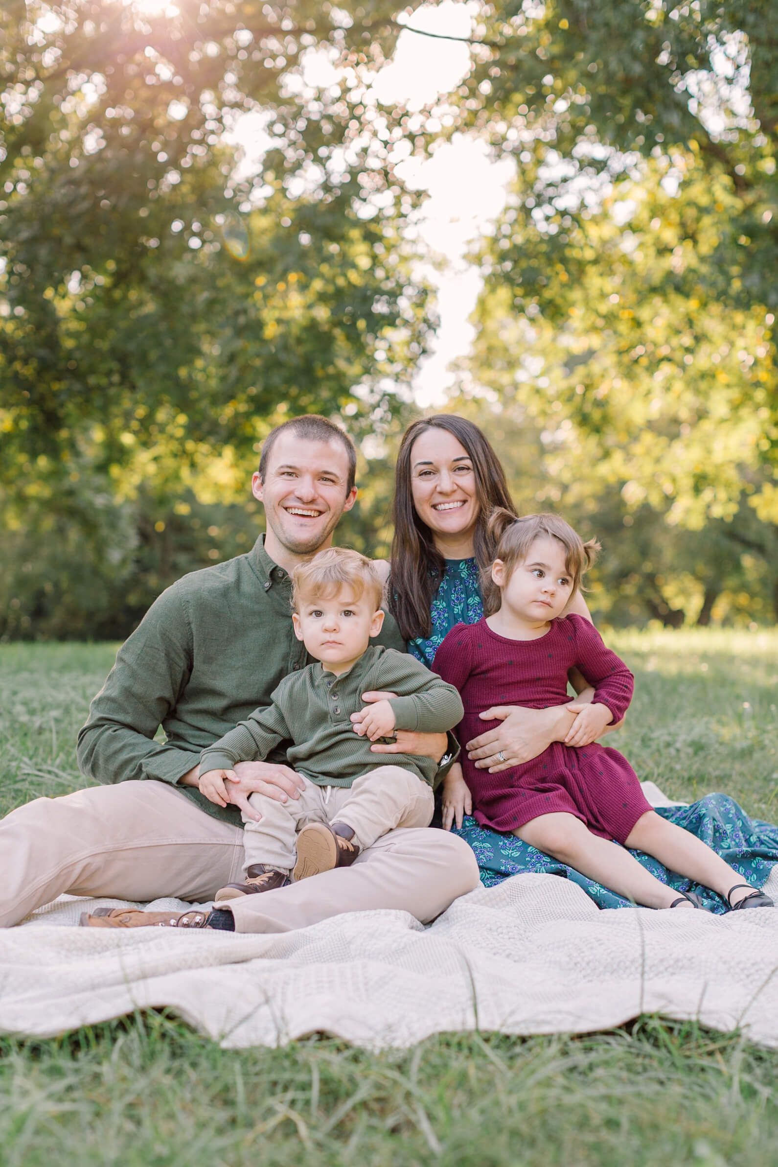 A happy mom and dad sit with their toddler son and daughter in their laps on a picnic blanket in a park at sunset