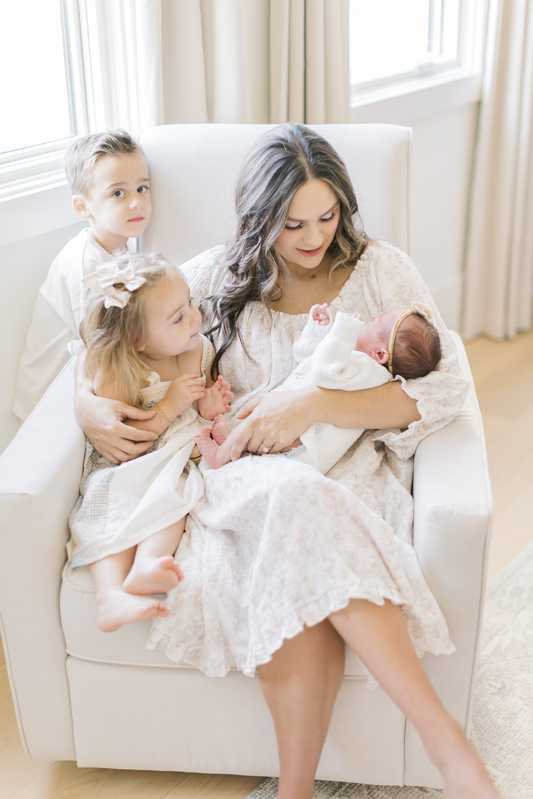A mother sits in a chair with her newborn and two toddlers under a window
