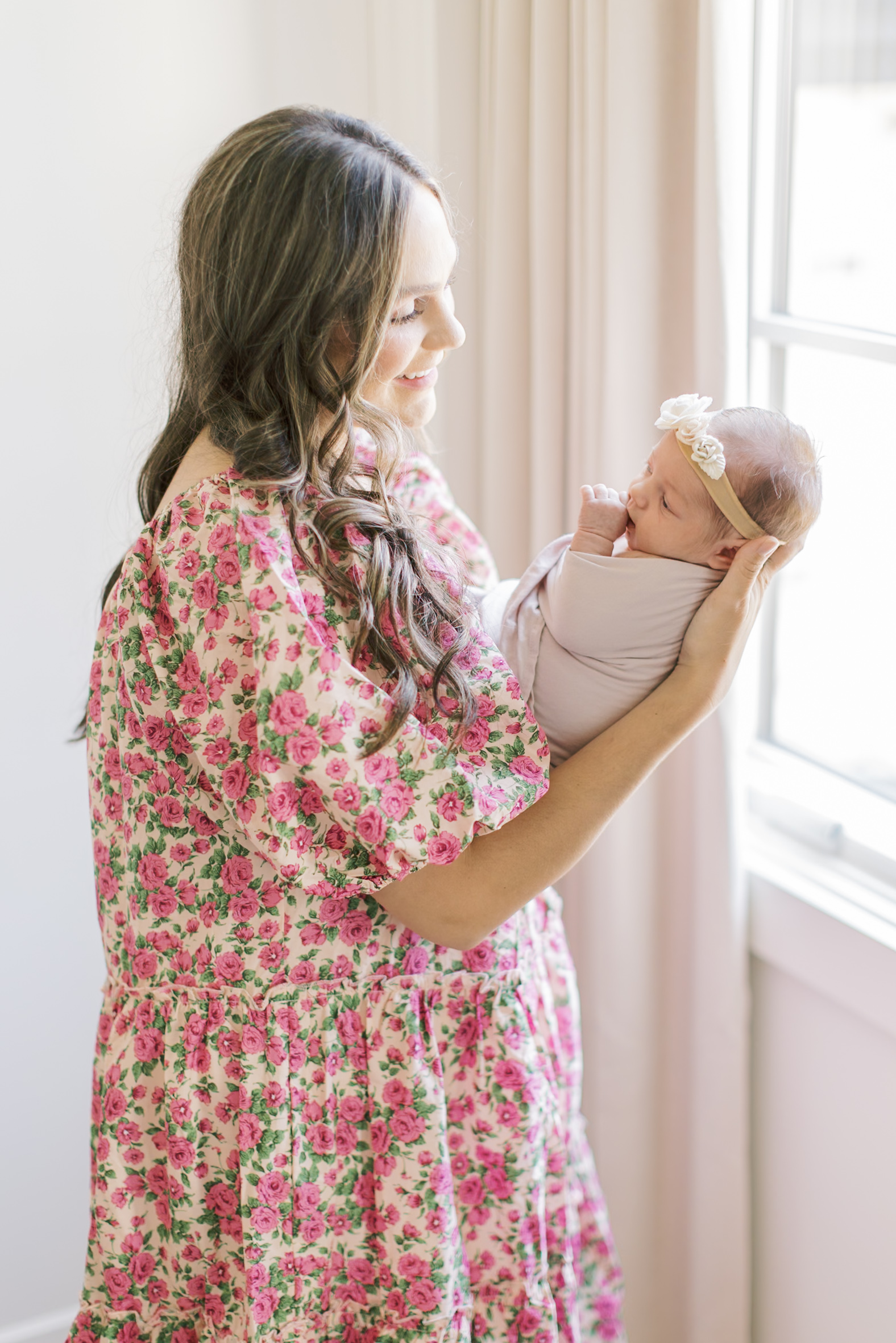 A happy mother in a pink flower print maternity dress stands in a window smiling down at her newborn before heading to Mother's day brunch in Raleigh, NC