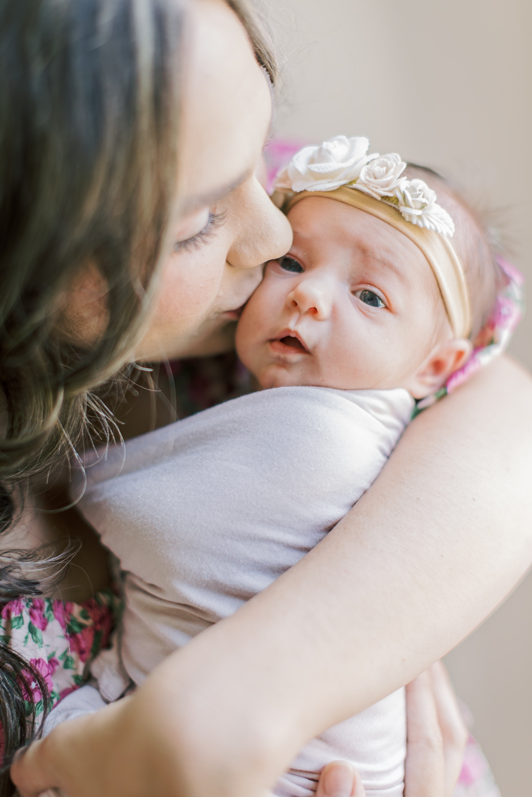 A mother kisses the cheek of her awake newborn while standing in a window waiting to head to Mother's day brunch in Raleigh, NC