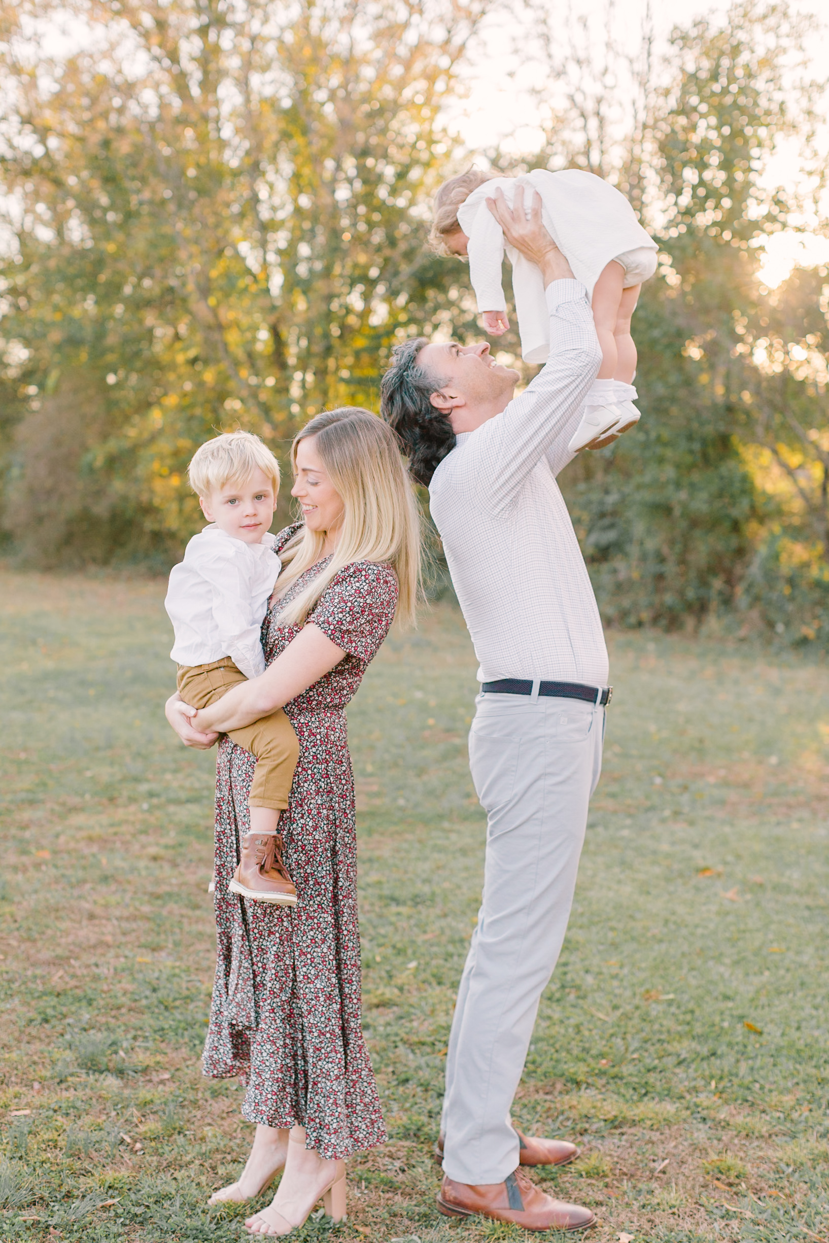 Happy mom and dad lift and play with their toddler son and daughter in a park lawn at sunset