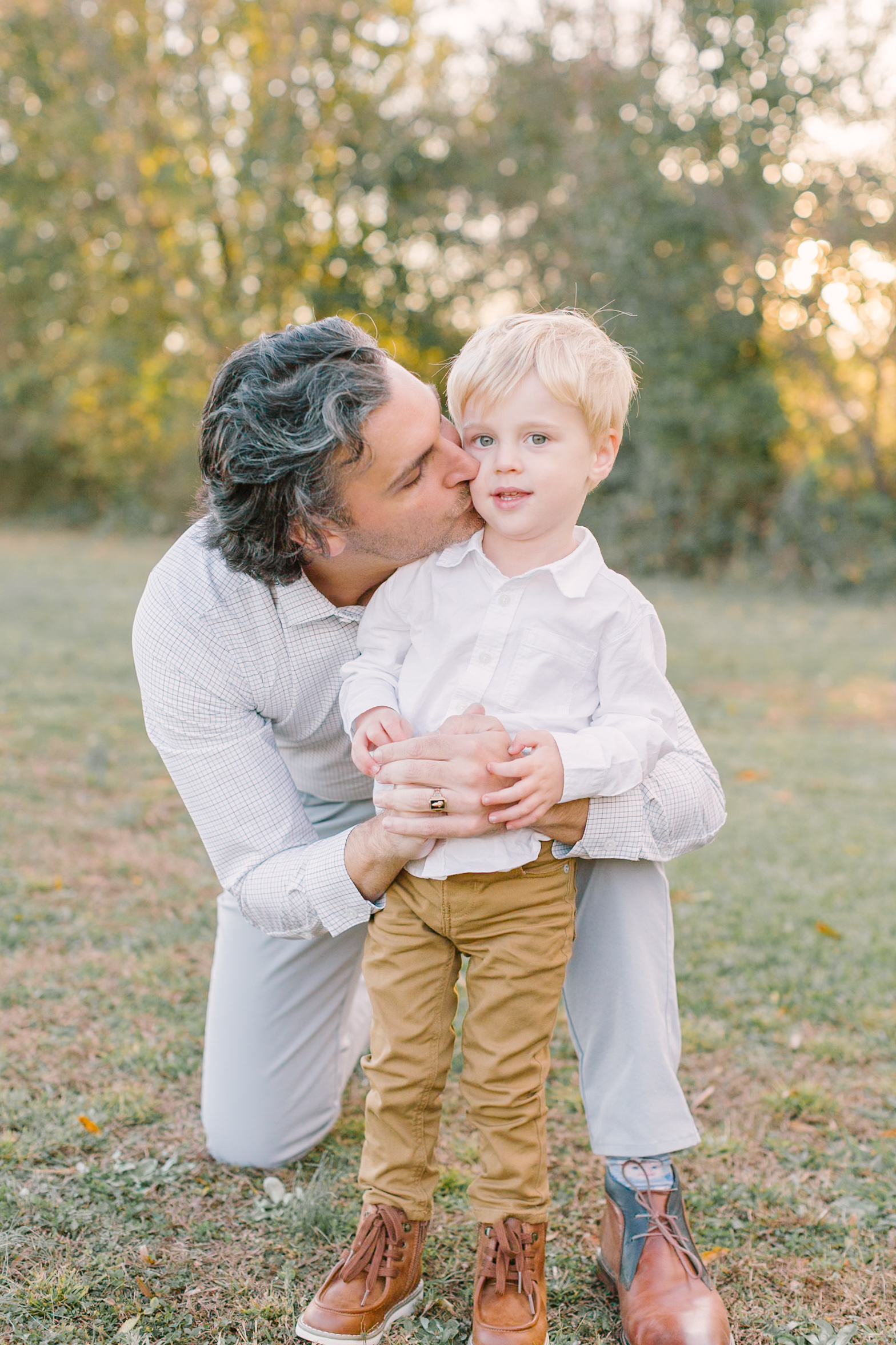 A dad kneels to kiss his toddler son on the cheek in a park at sunset
