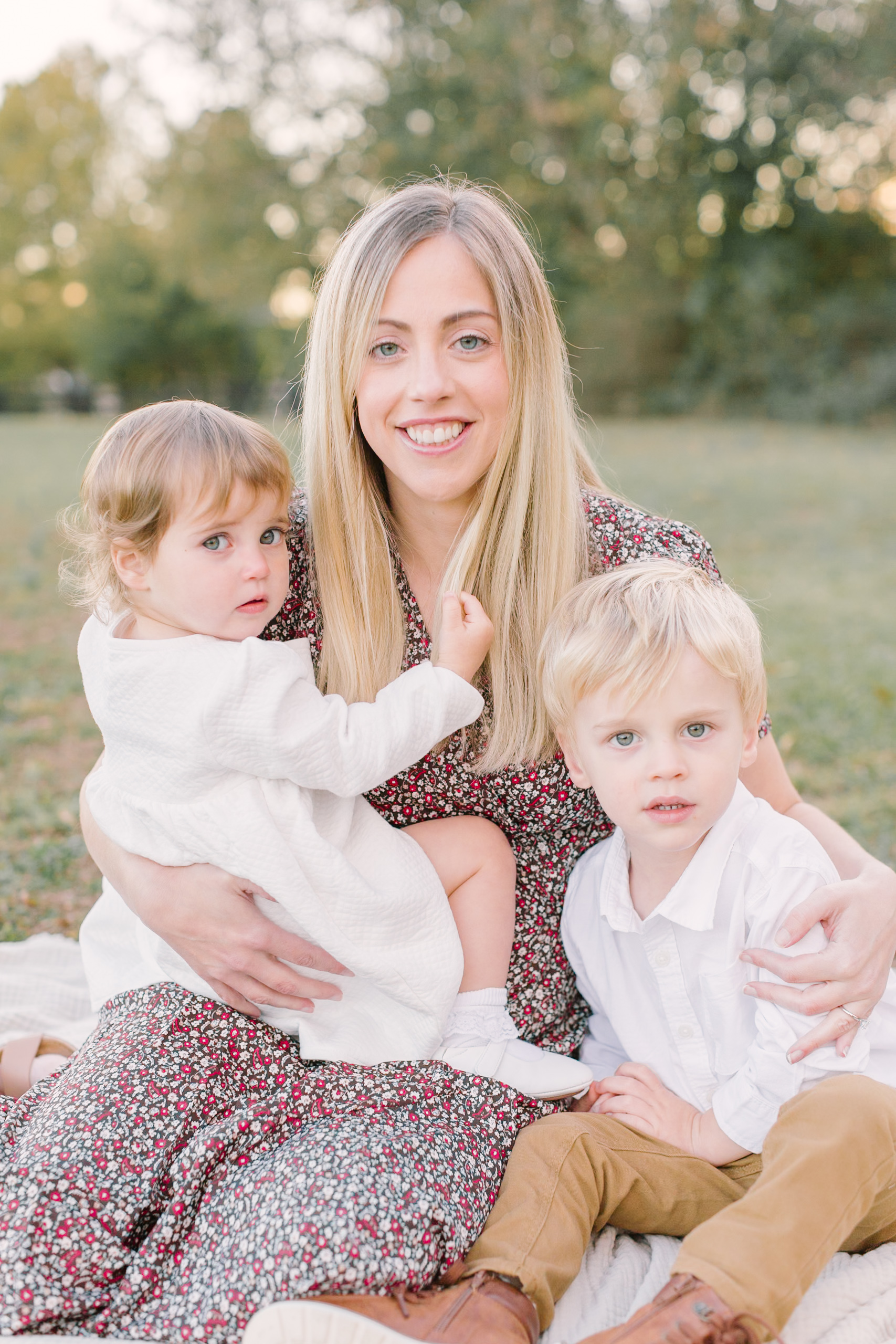 A happy mom in a floral print dress hugs her toddler son and daughter on a picnic blanket at sunset after finding their easter sunrise services in Raleigh, NC