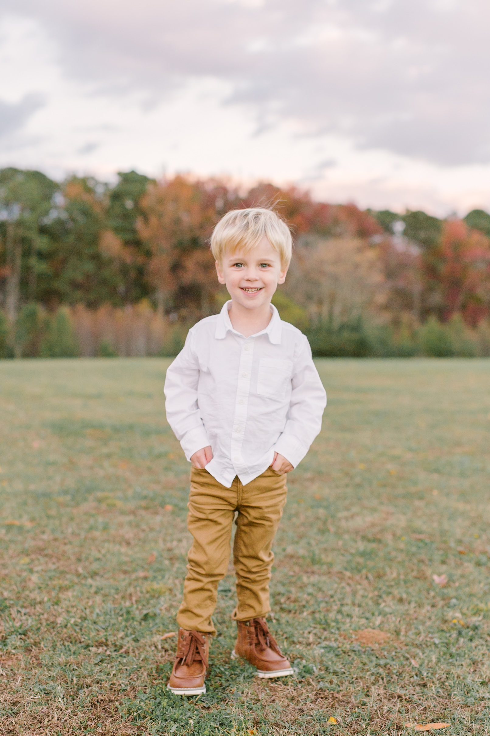 A toddler boy in a white shirt and khakis stands smiling in a field at sunset before exploring easter sunrise services in Raleigh, NC