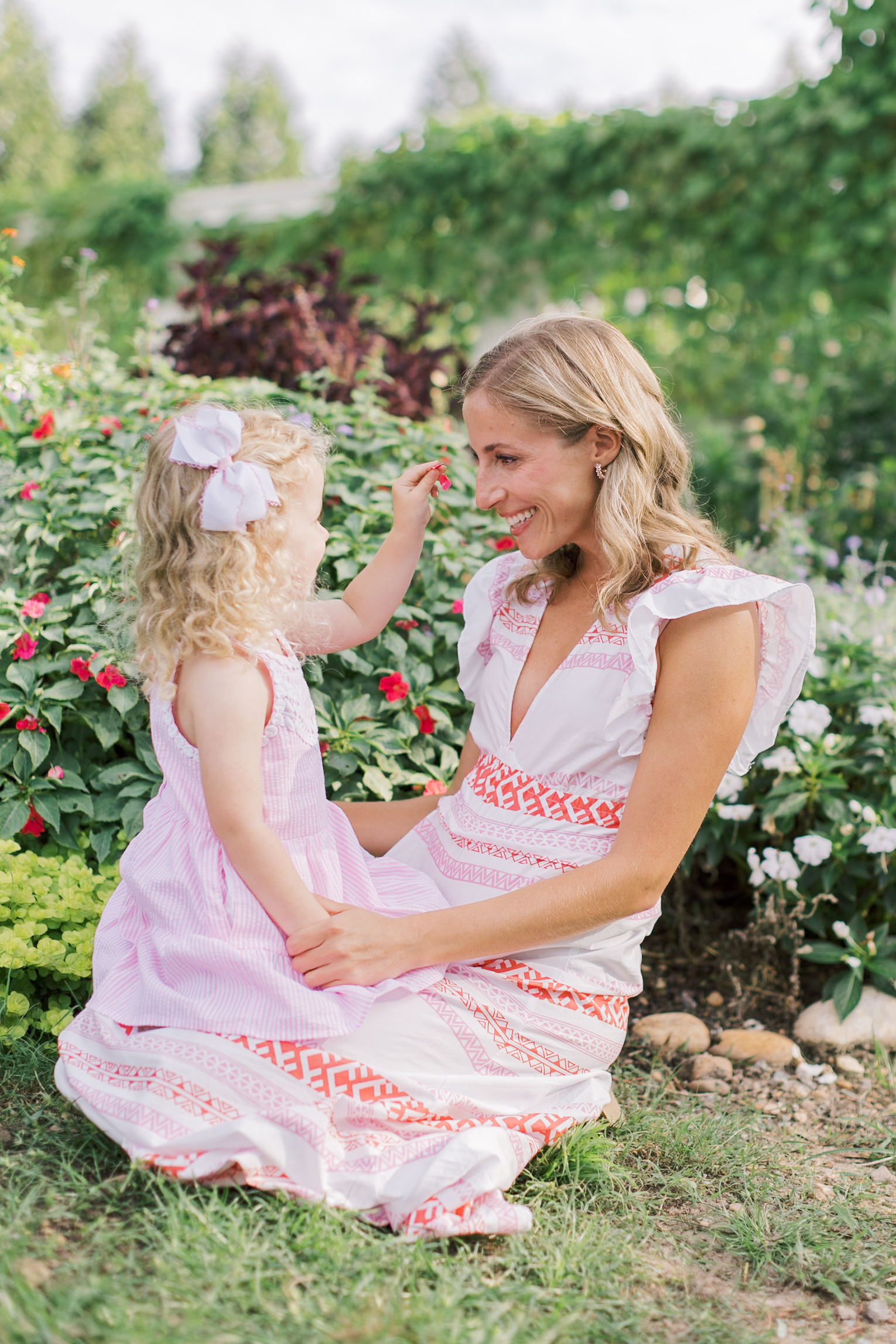a toddler girl in a pink dress sits in mom's lap surrounded by flowers playing and laughing