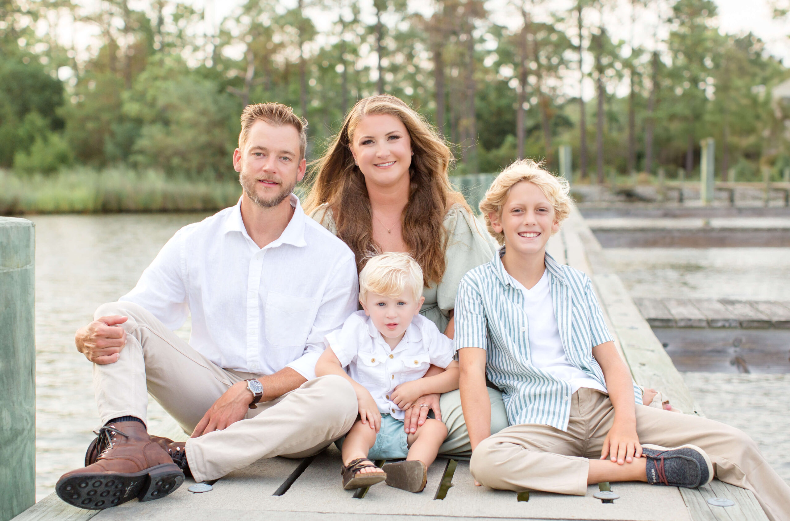 A happy mom and dad sit on a lake dock with.their two young sons in their laps