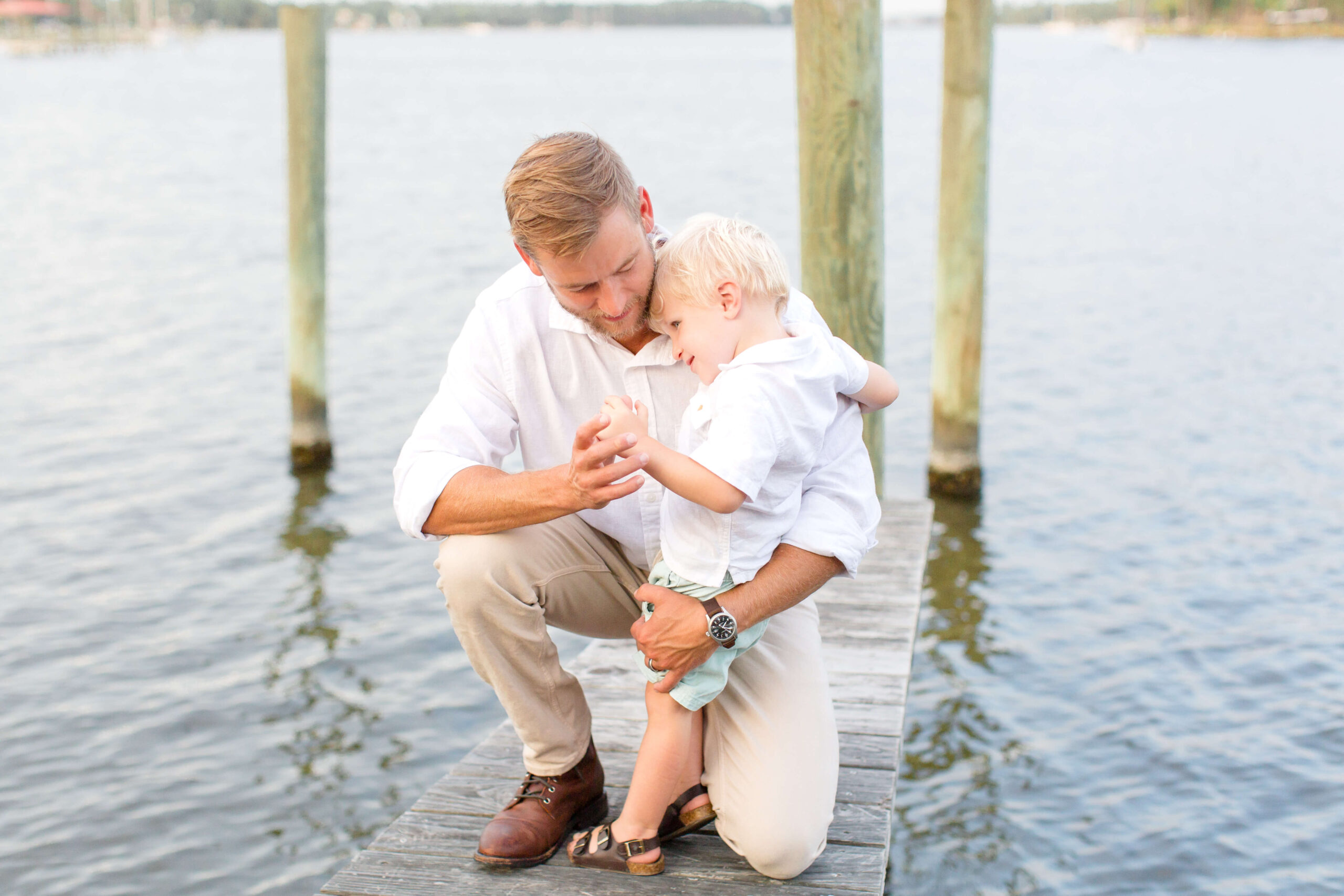 A father plays with his toddler son on a skinny boat dock at sunset in white shirts