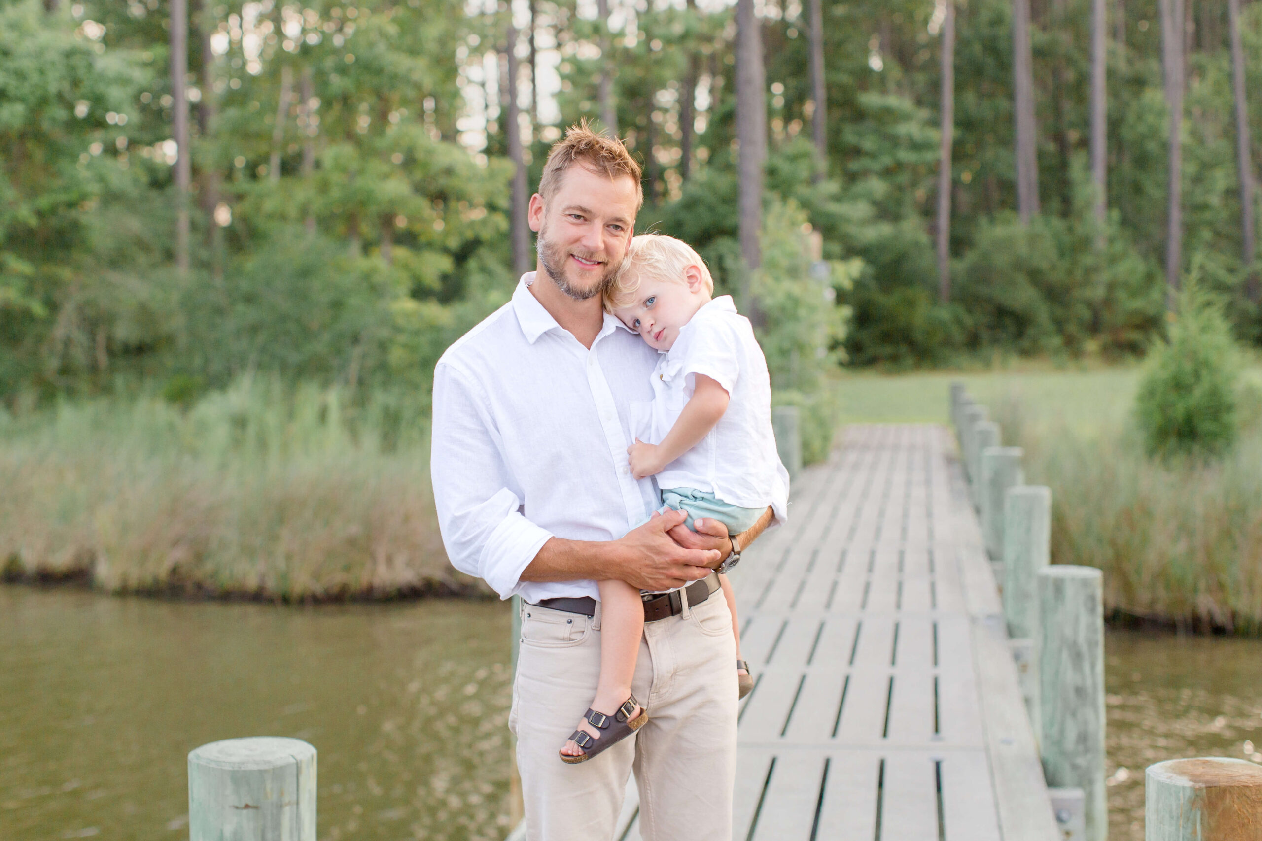 A happy dad walks on a lake dock holding his tired toddler son both in white shirts after enjoying some outdoor dining in Raleigh, NC