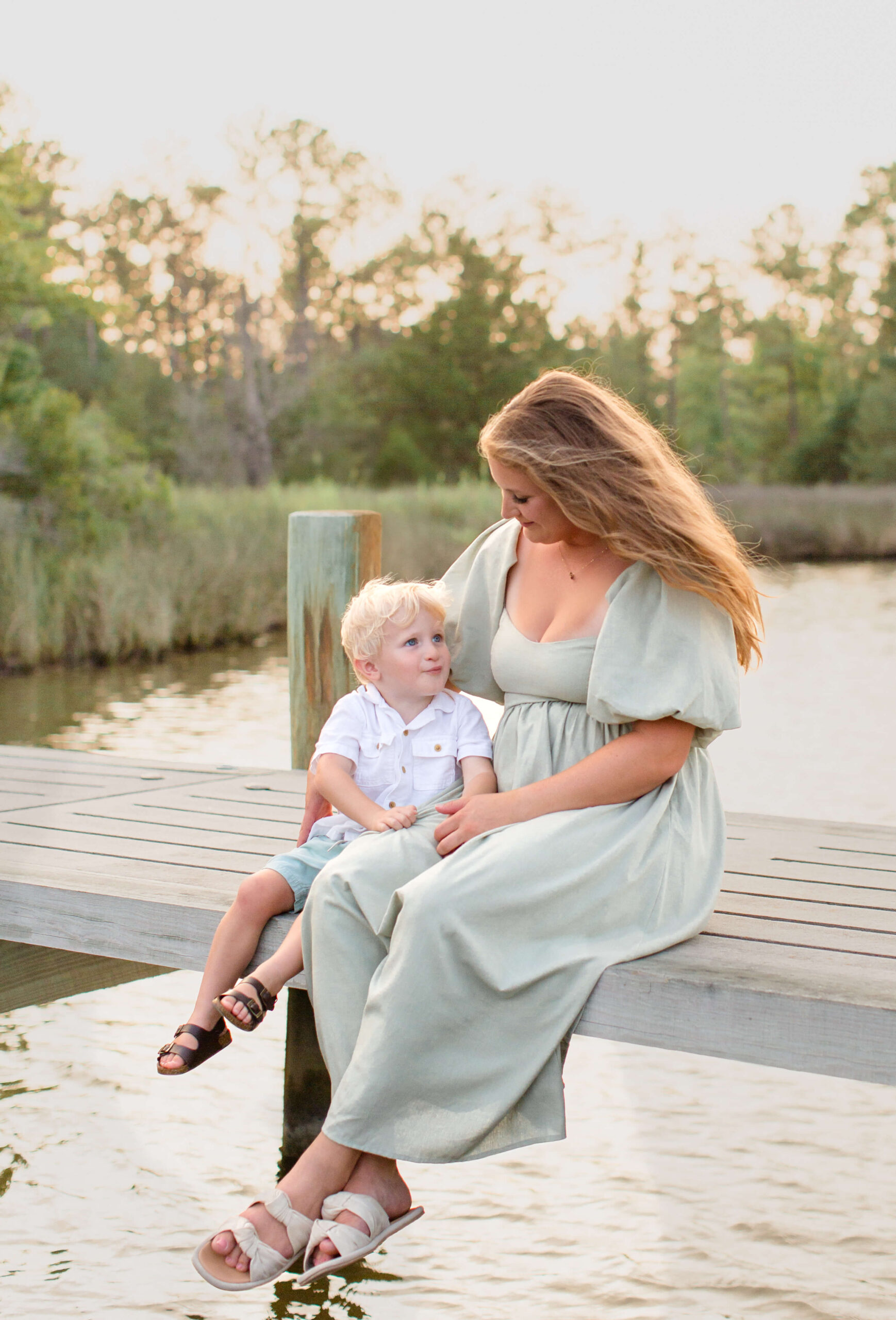 A mother in a green dress sits on a dock at sunset smiling and cuddling her toddler son after enjoying outdoor dining in Raleigh, NC