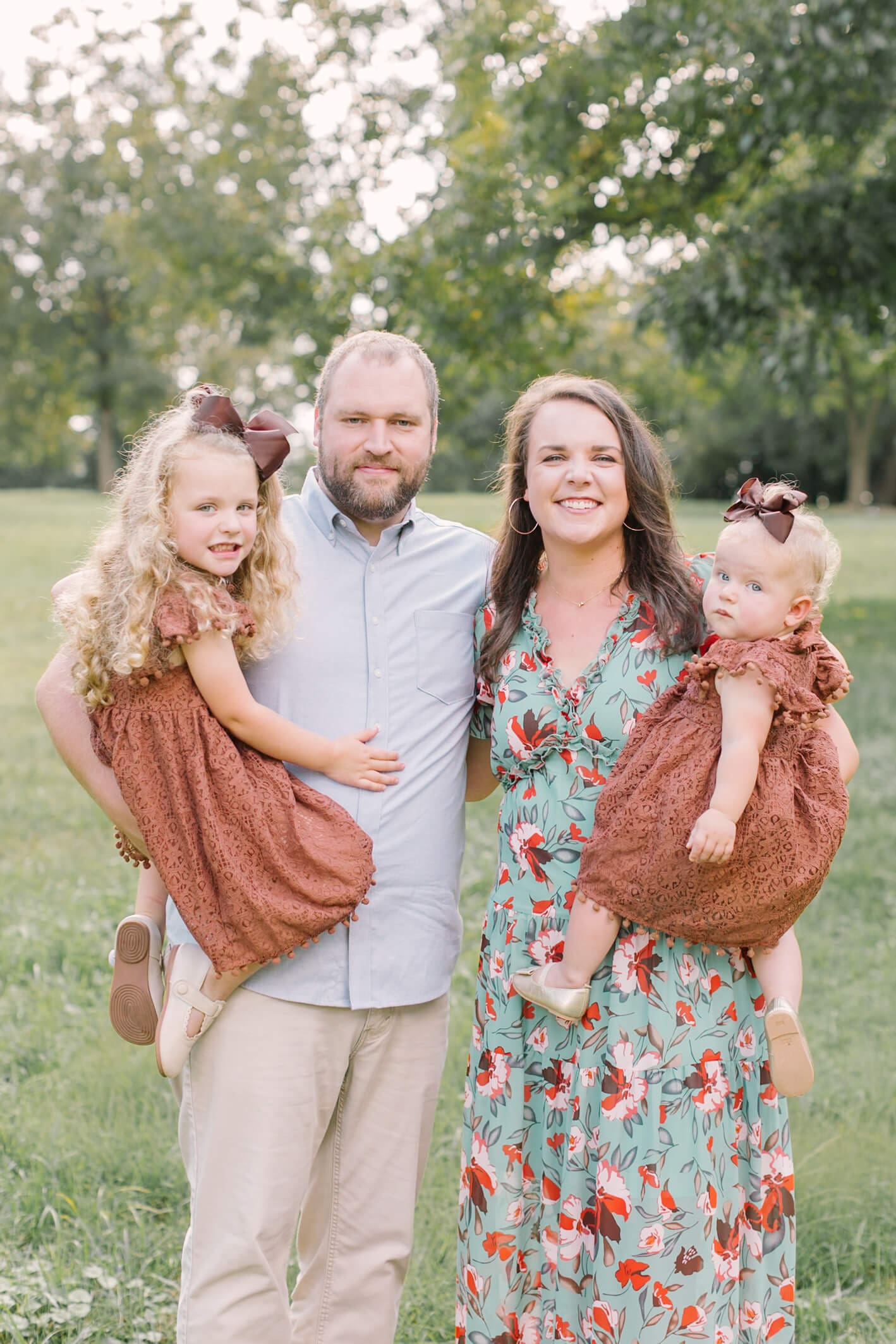 A smiling mom and dad stand in a park lawn holding their two toddler daughters on their hips
