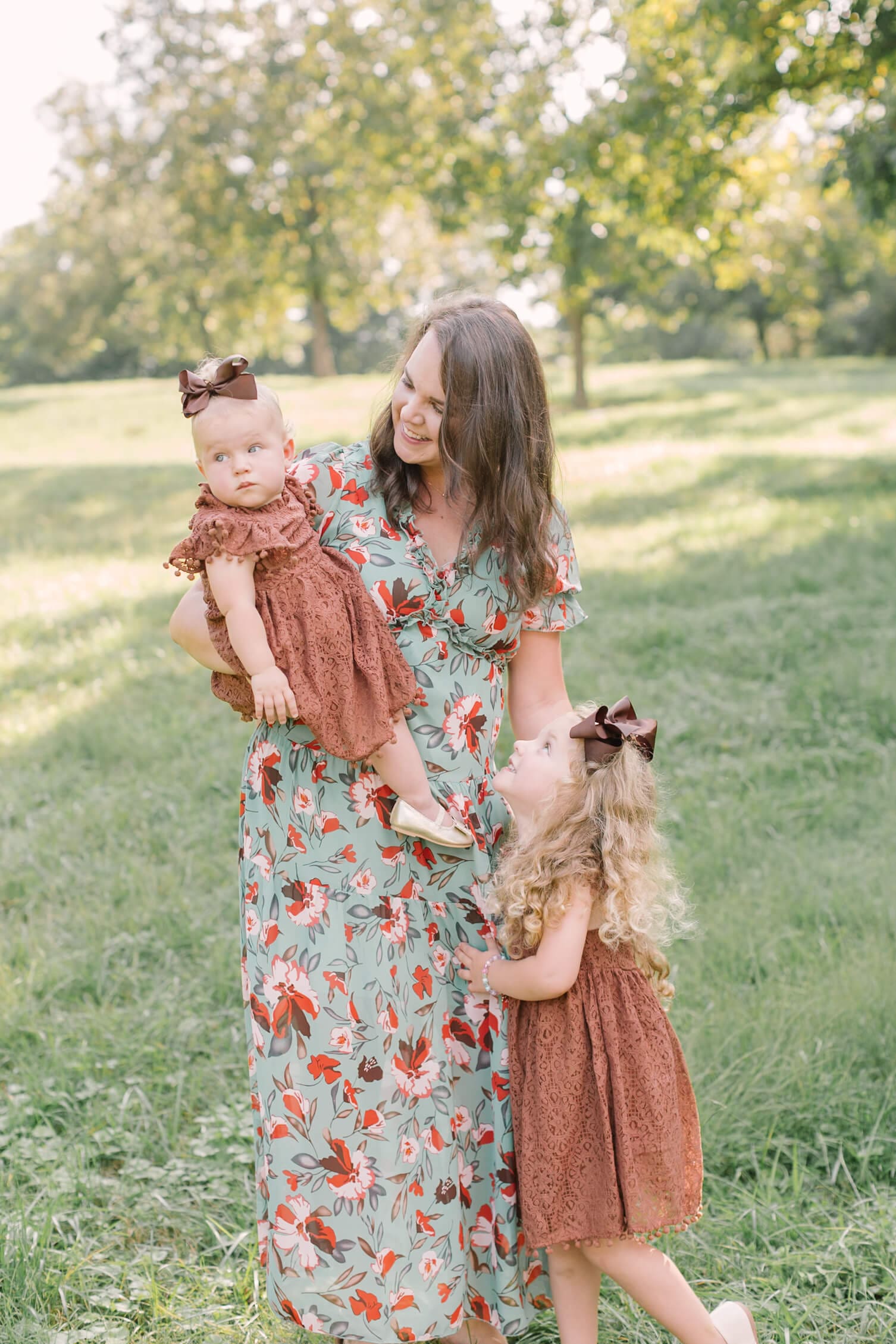 A happy mom in a green floral print dress plays win a park with her two toddler daughters in matching brown dresses and bows