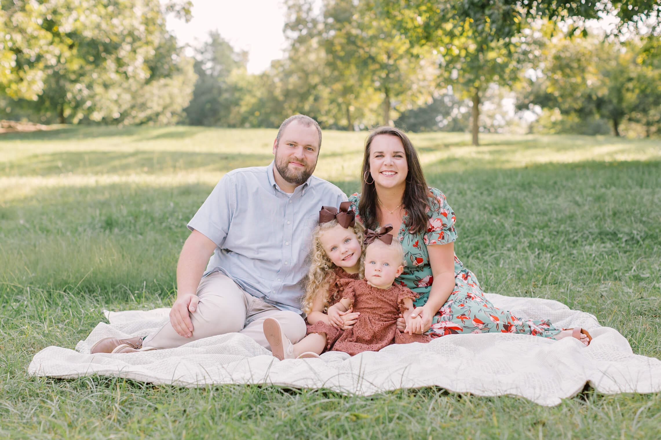Happy mom and dad sit on a picnic blanket in a park lawn with their two toddler daughters in matching brown dresses before some strawberry picking in raleigh, nc