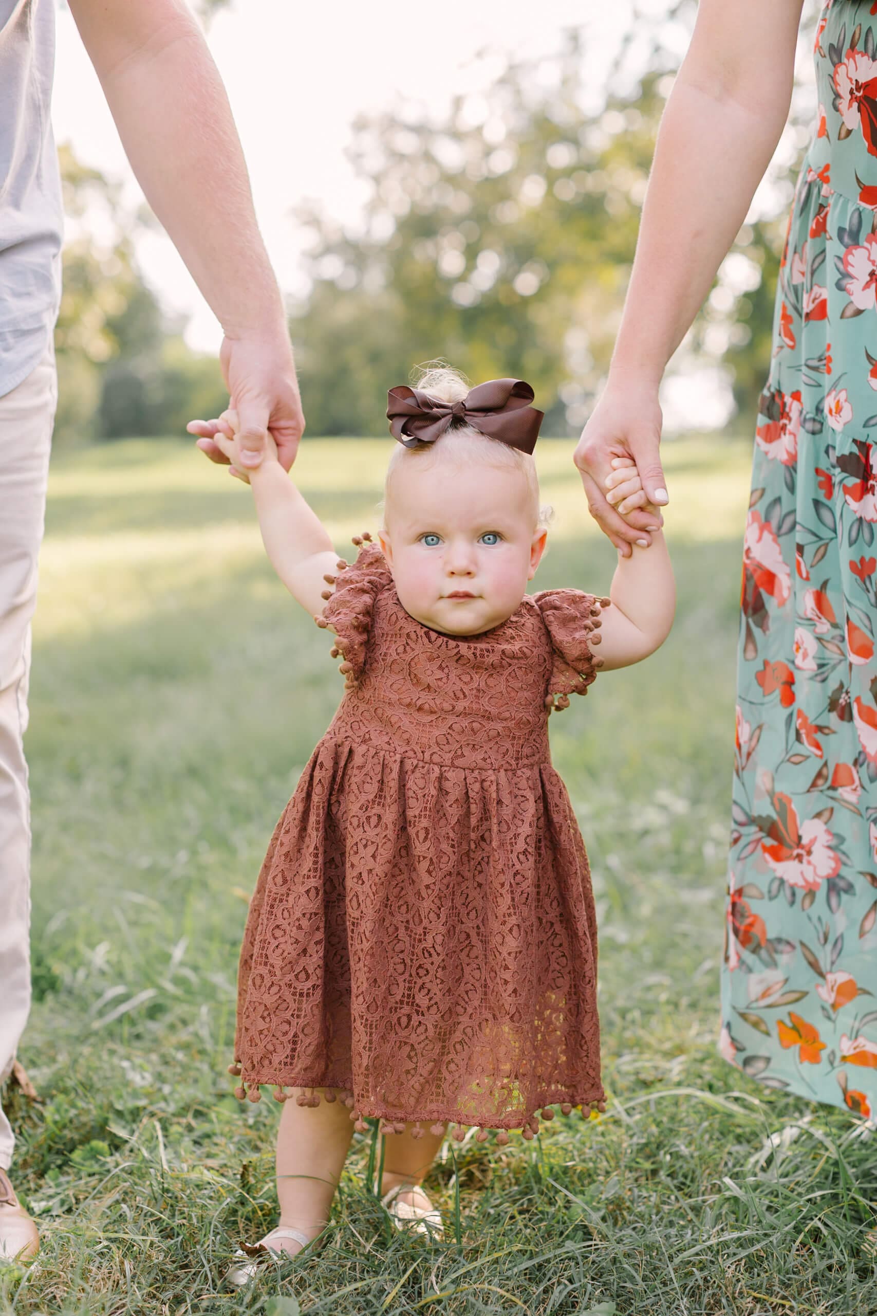 A toddler girl in a brown lace dress walking in a park lawn holding mom and dad's hands before some strawberry picking in raleigh, nc