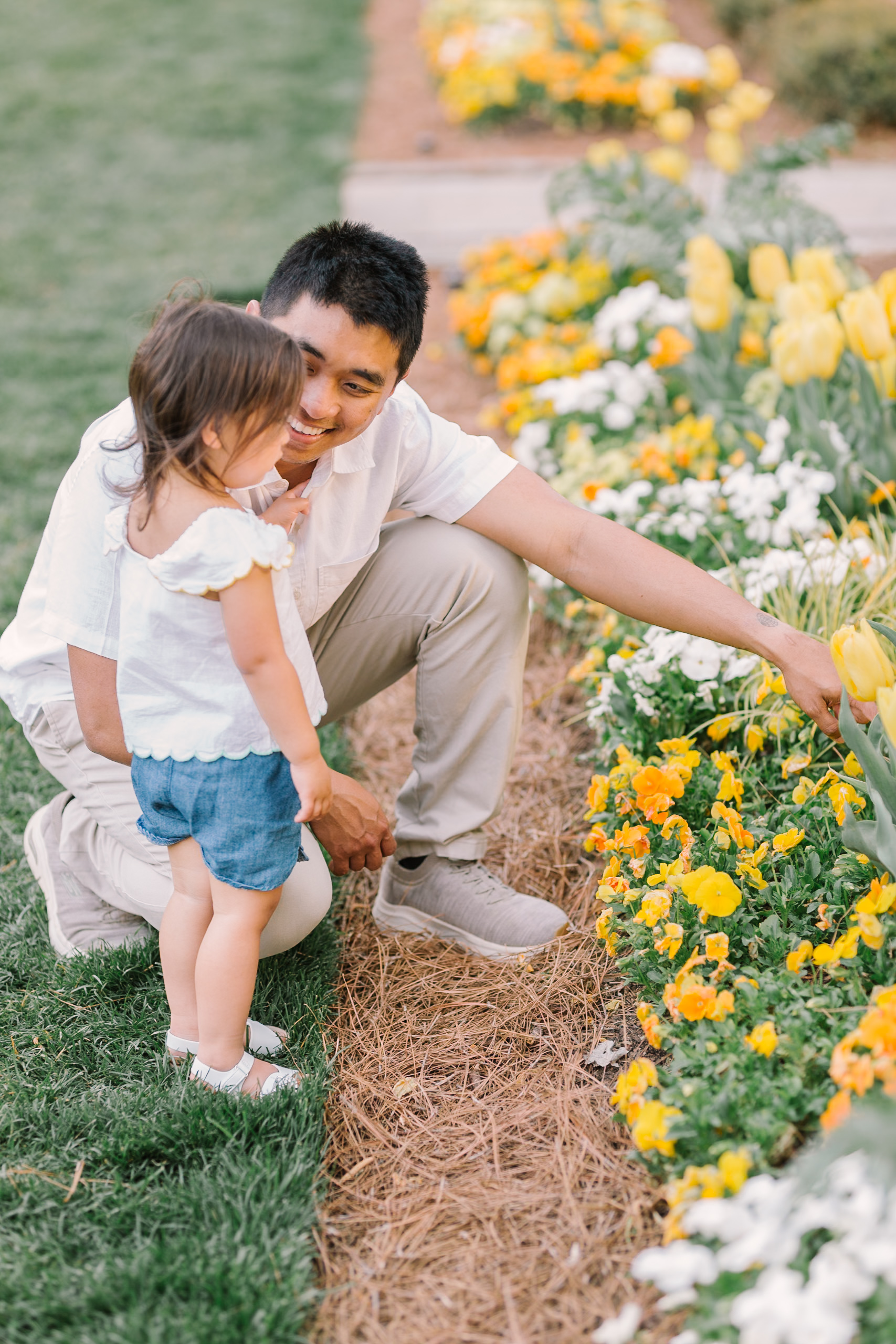 A dad in white and khakis points to some flowers in a garden at sunset to his toddler daughter in white and denim