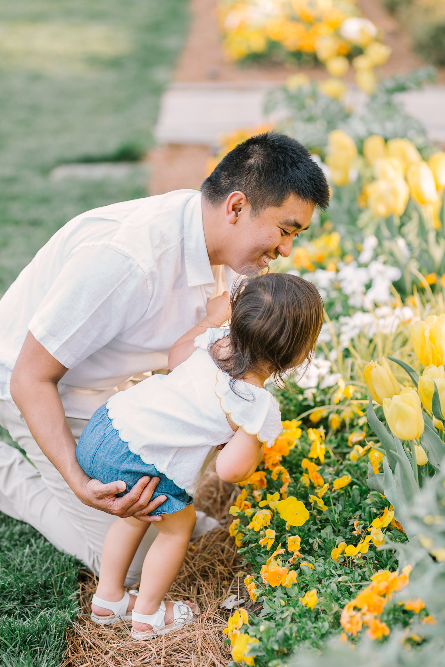 A happy dad shows his toddler daughter some yellow flowers in a garden in white shirts