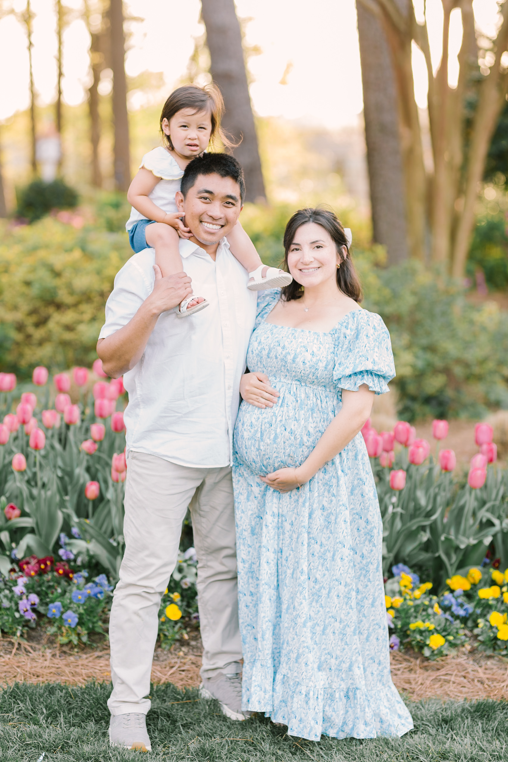 Happy pregnant mom and dad stand in front of one of the flower fields in Raleigh, NC holding the bump in blue and white with toddler on dad's shoulders