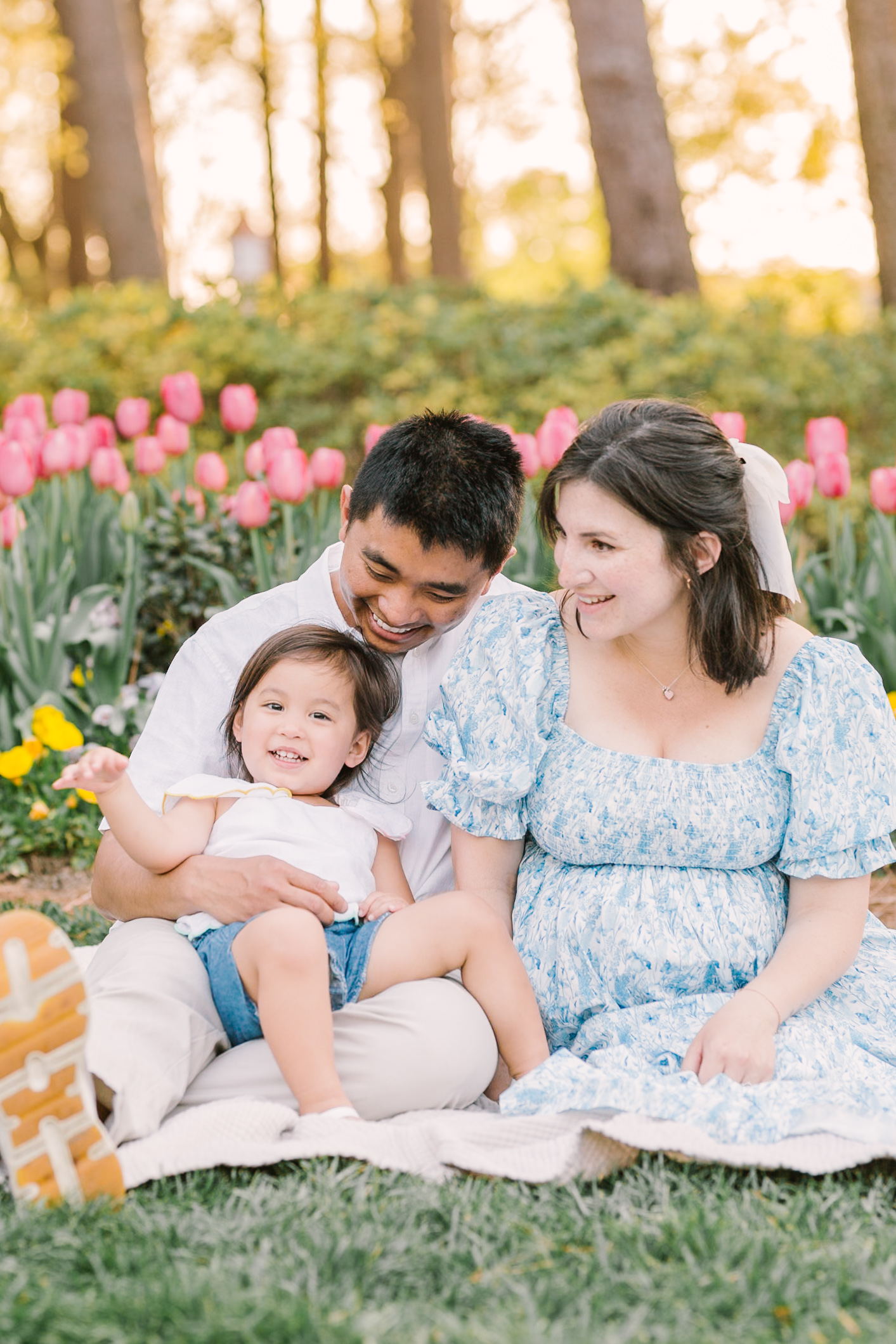 Happy mom and dad sit in a tulip flower field in Raleigh, NC on a picnic blanket with their toddler daughter in their laps