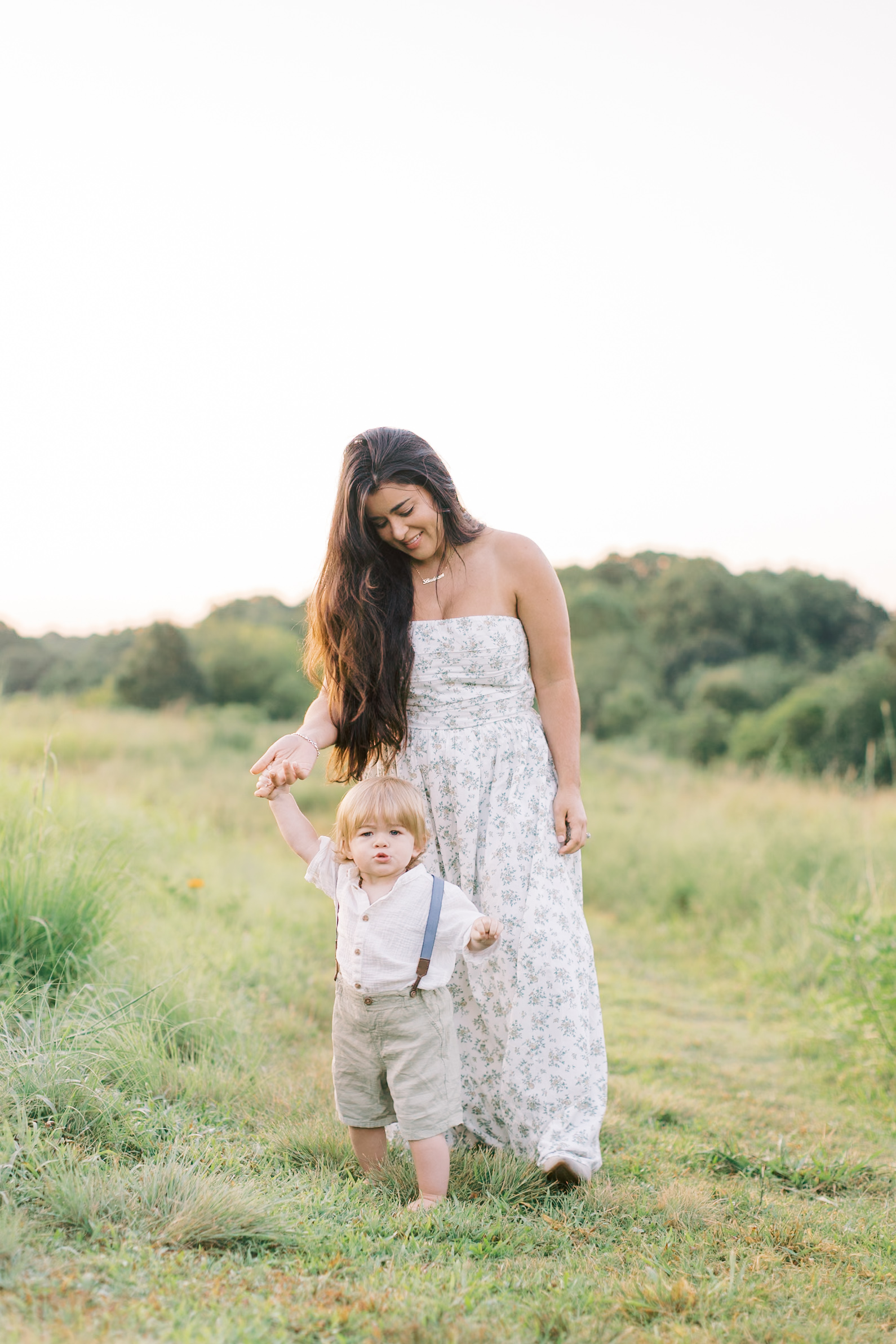 A mother wearing a long floral dress smiles and looks down at her baby as they walk in a field at sunset.
