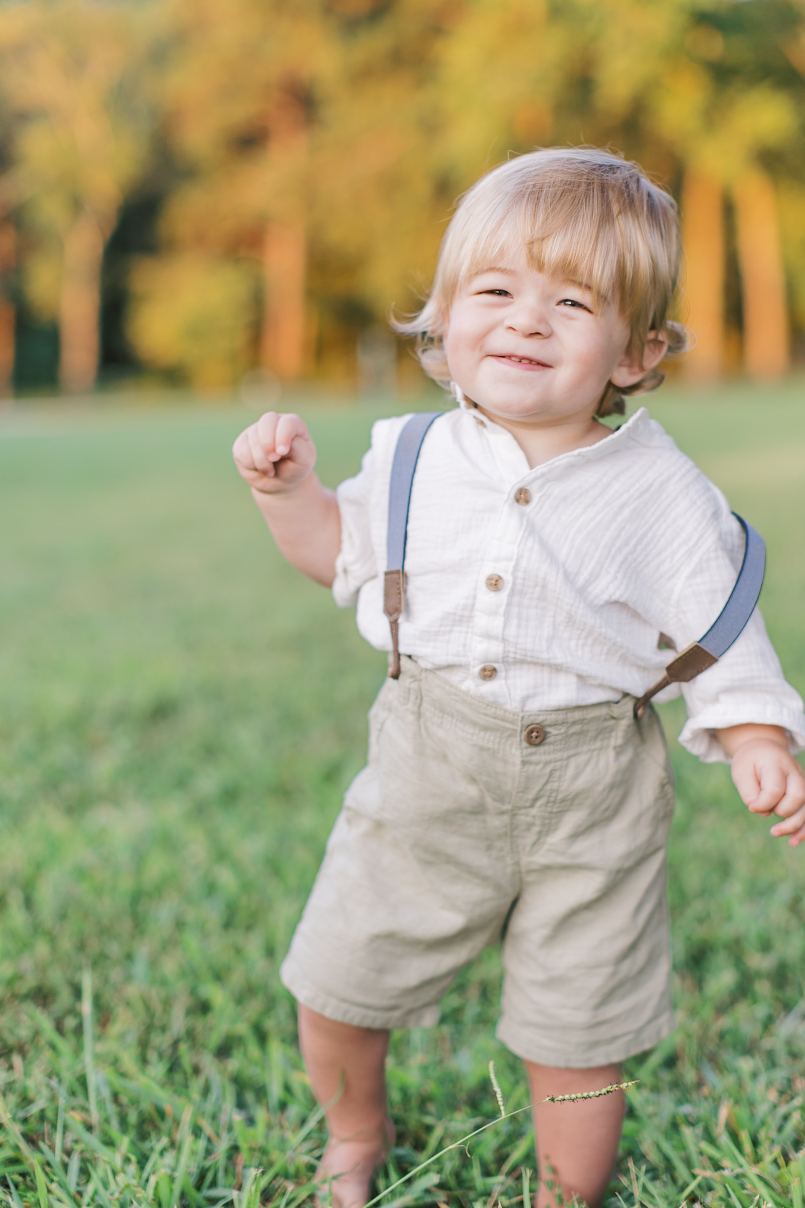 A blonde baby waddles in a patch of grass as he smiles while wearing a white button up and beige pants with blue suspenders holding them up.