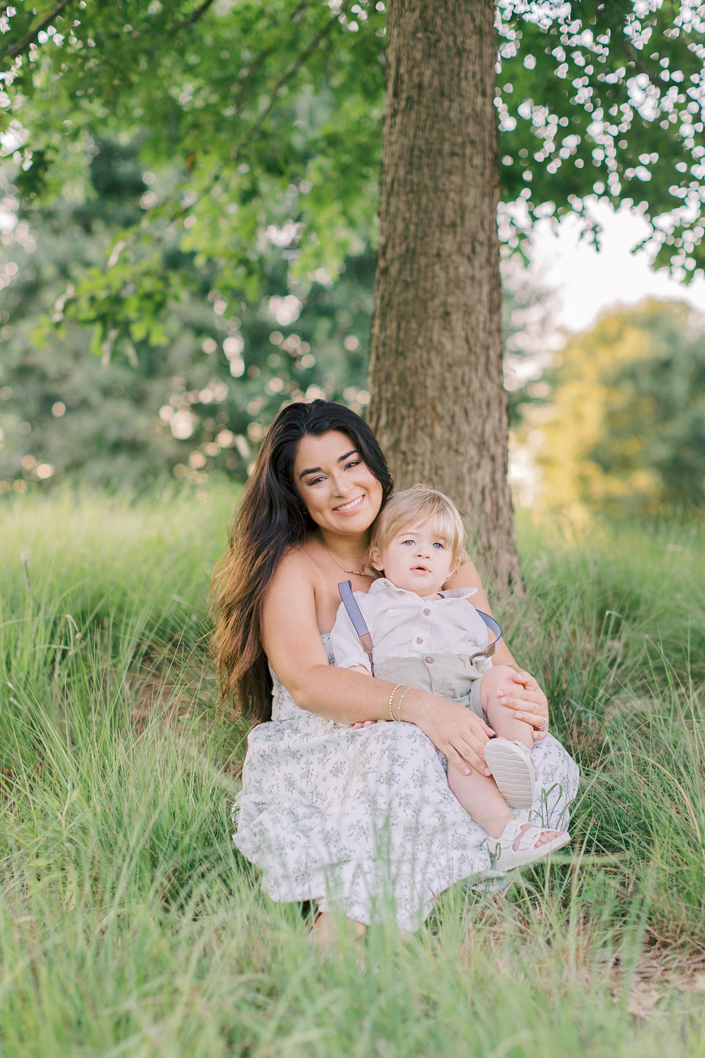 A mother sits in a patch of grass in front of a tree as she holds her young son who is sitting on her lap after enjoying baby music classes in raleigh nc.
