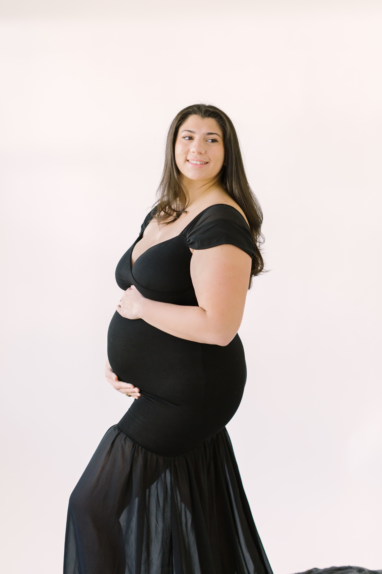 A smiling mother to be holds her bump while standing in a studio looking over her shoulder in a black maternity gown