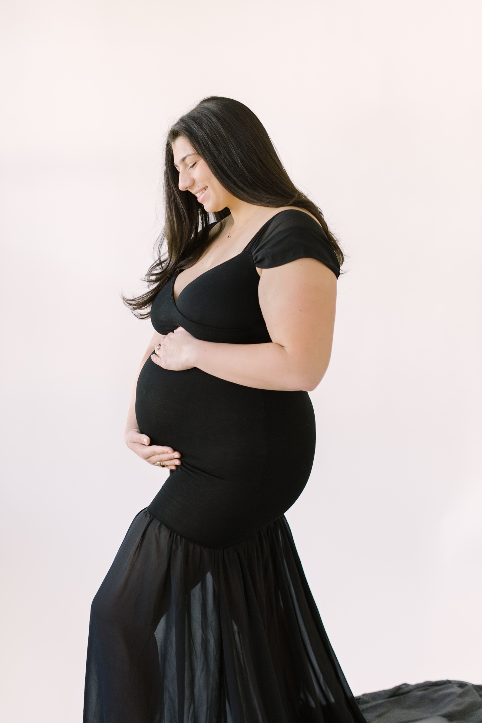 A happy mom to be smiles down to her bump with hands on top and bottom in black while standing