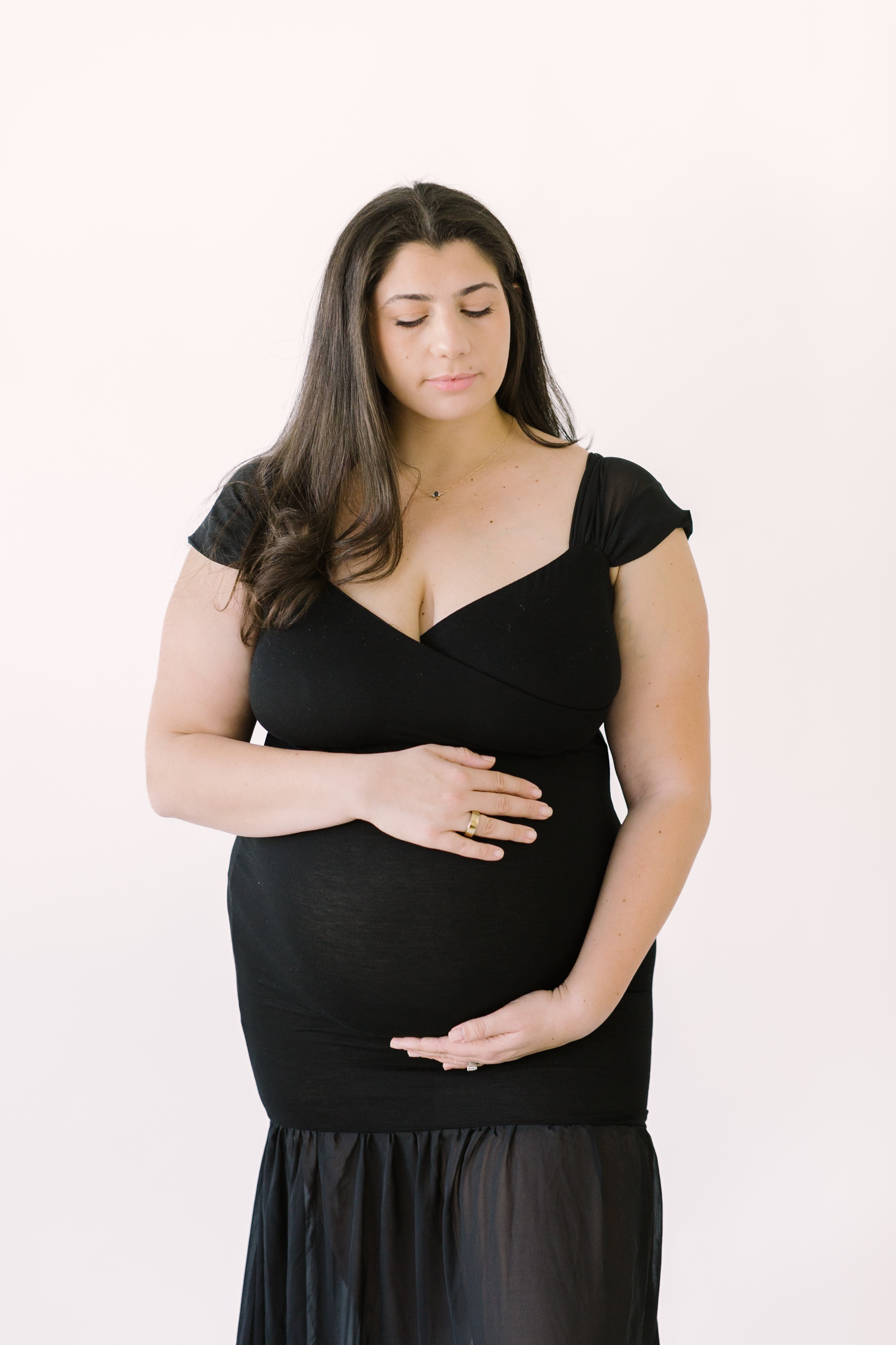 A mother to be in a black maternity gown stands with hands on her bump in a studio thanks to fertility clinics in Raleigh, NC