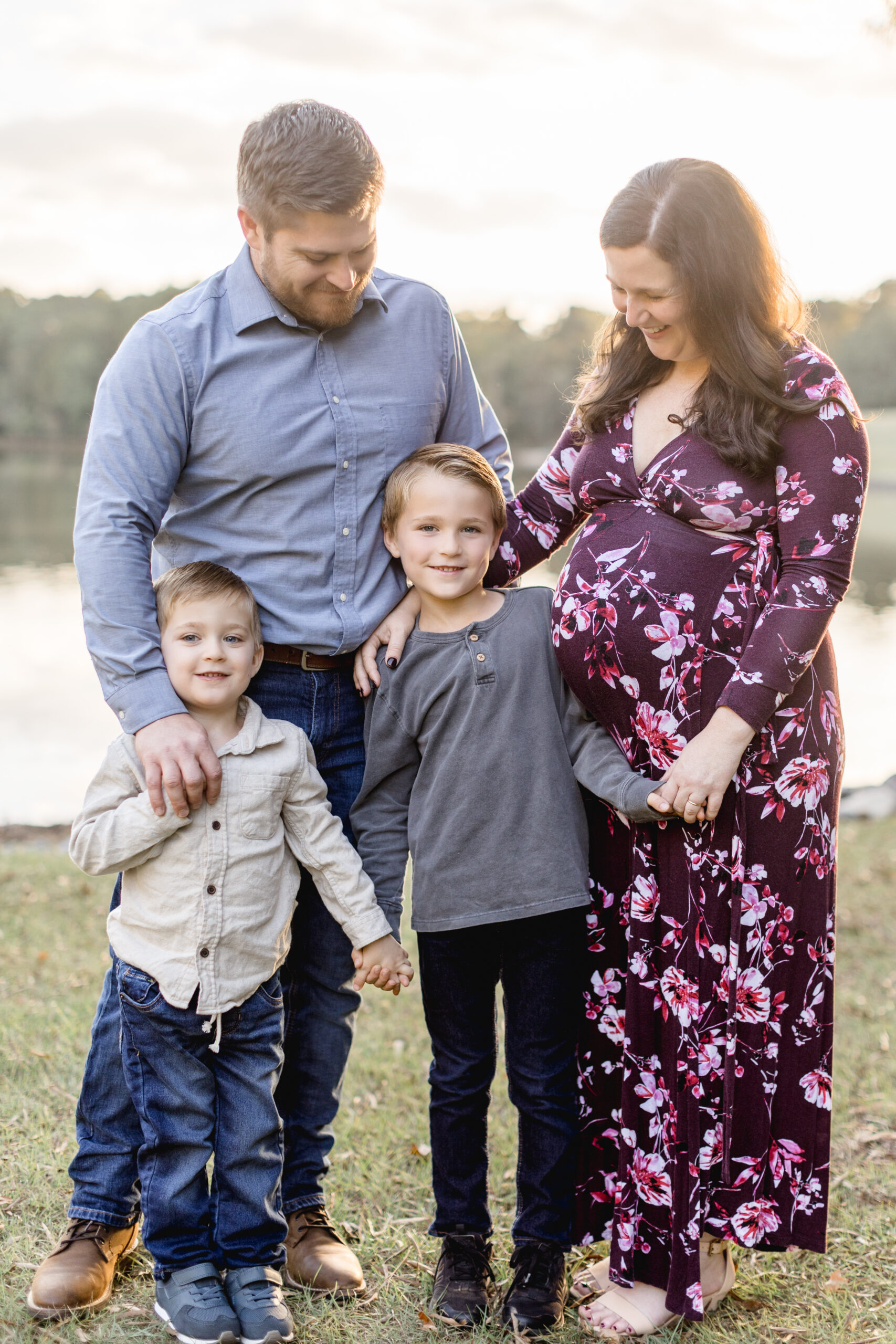 A couple smiles down on their two sons who are holding hands and smiling at the camera as they stand in front of a pond.