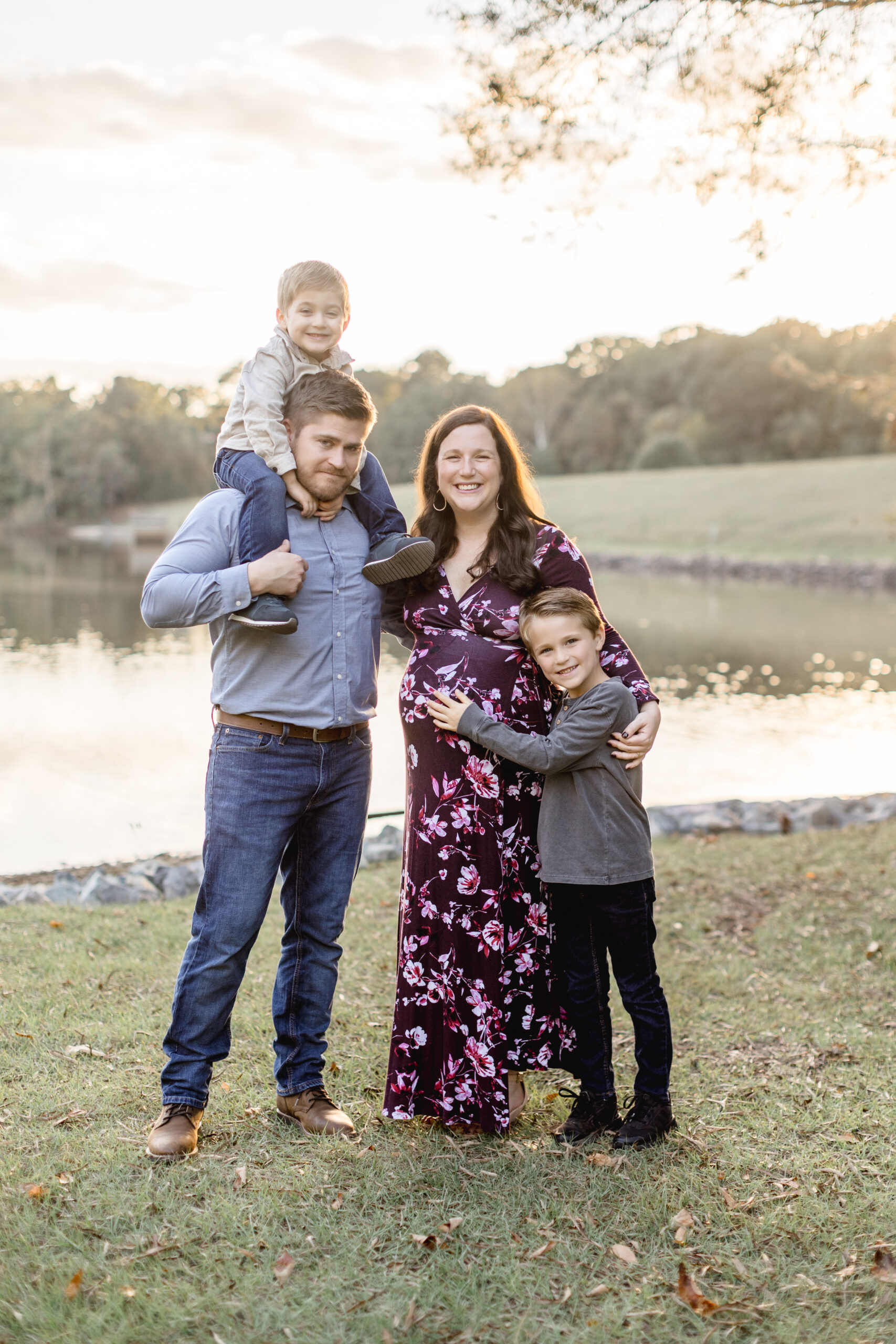 A family stands, smiling in front of a pond at sunset while the dad holds one son on his shoulders and the other son hugs his mothers pregnant bump.