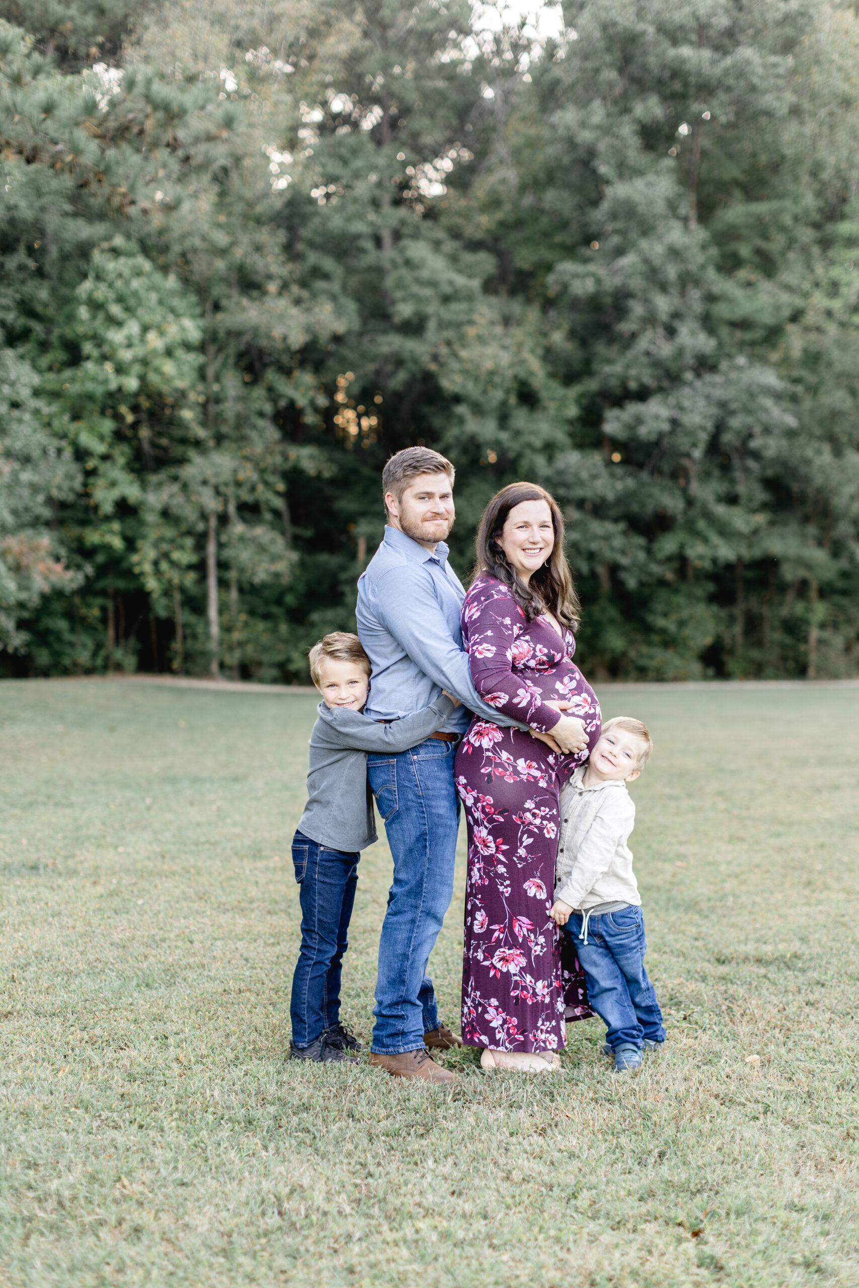 A family stands in a park as the father holds his pregnant wife's bump while their two younger sons stand beside them and hugs them after visiting prenatal chiropractors in raleigh nc.