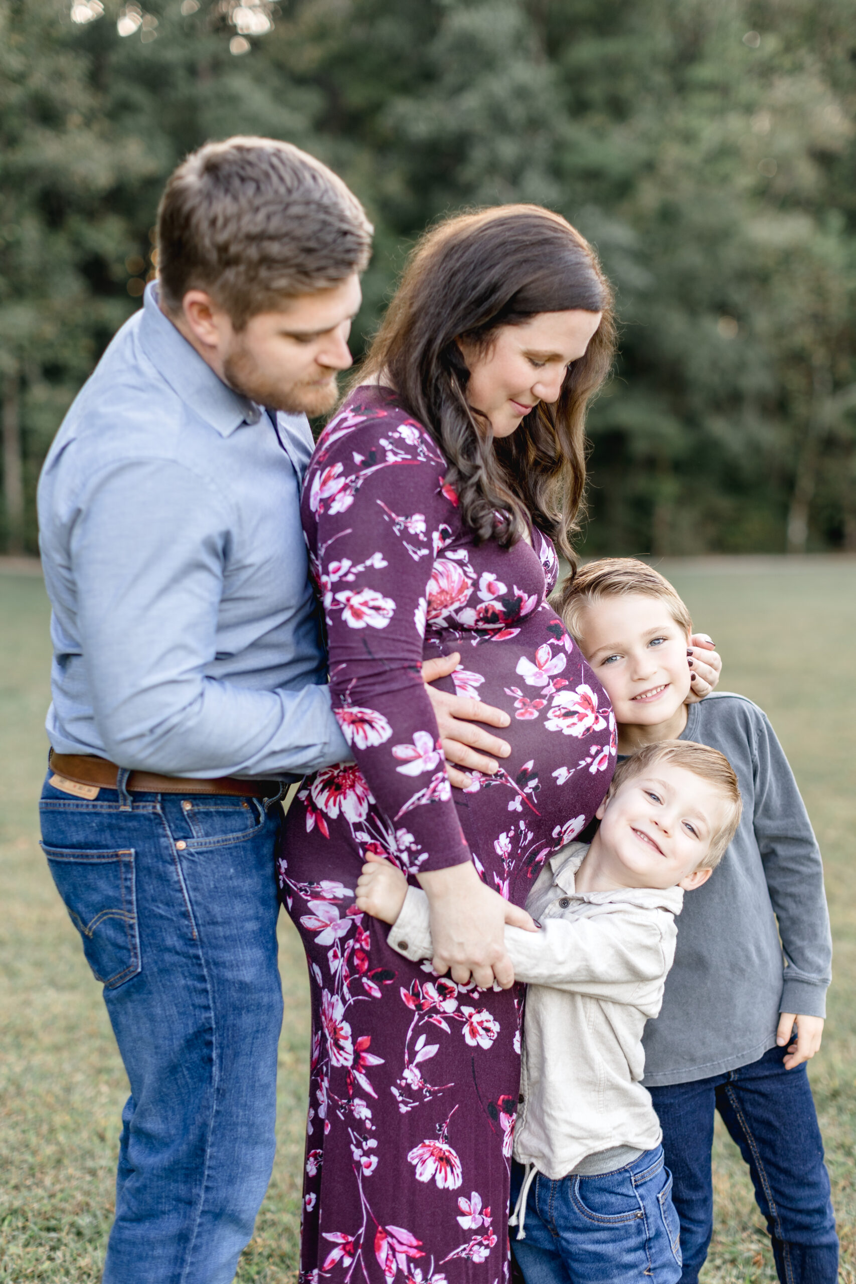 A father stands behind his pregnant wife and looks at his sons as he holds her bump while their two sons smile and hug the moms bump after seeing prenatal chiropractors in raleigh nc.
