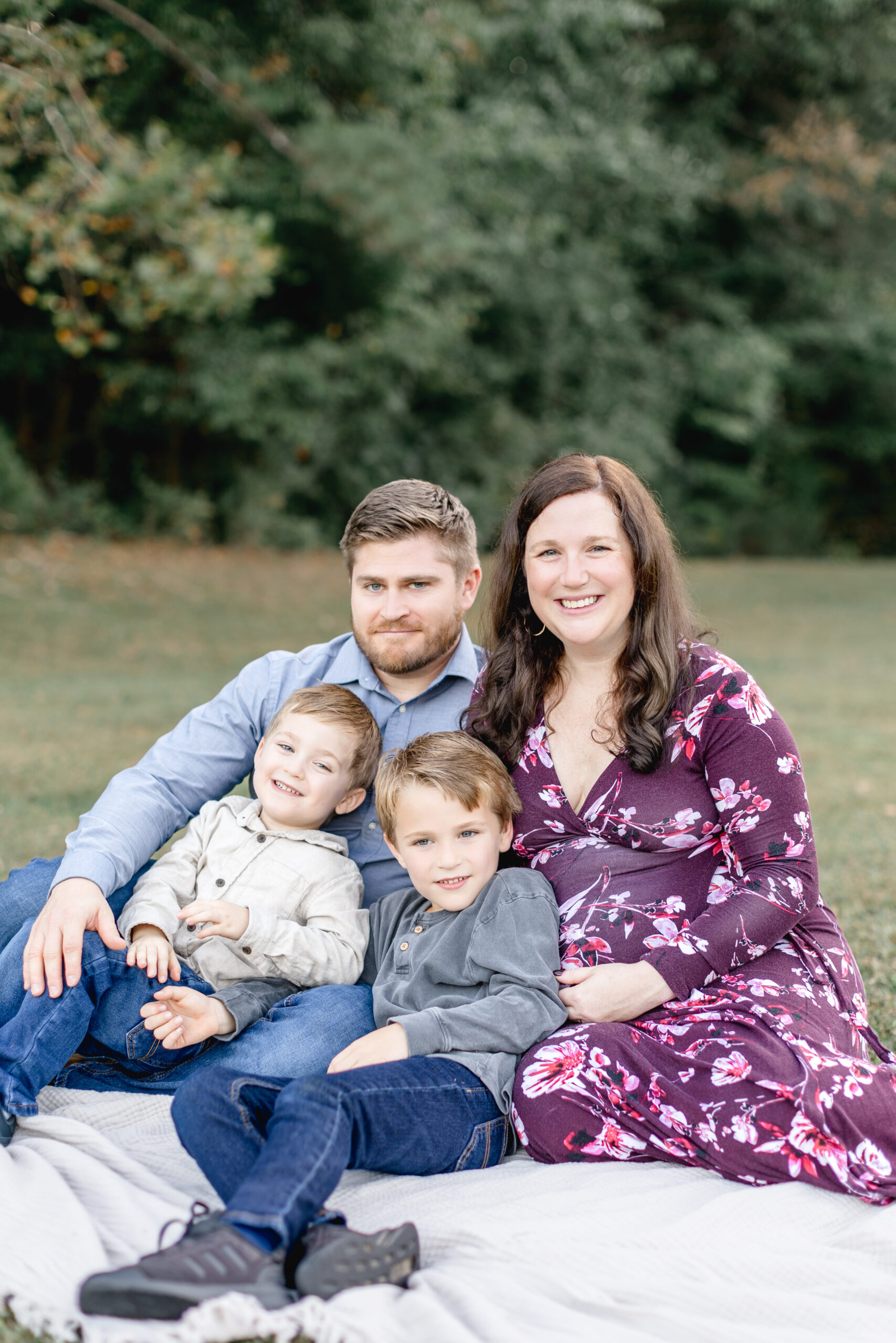 A happy couple and their two smiling toddler sons sit on a blanket in a park after finding a prenatal chiropractors in raleigh nc.